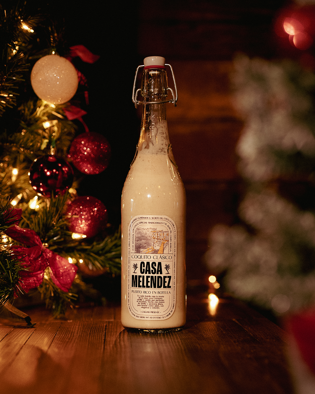 A bottle of Casa Melendez Puerto Rican Coquito Clásico on a wooden surface with a Christmas tree decorated with lights and red ornaments in the background.