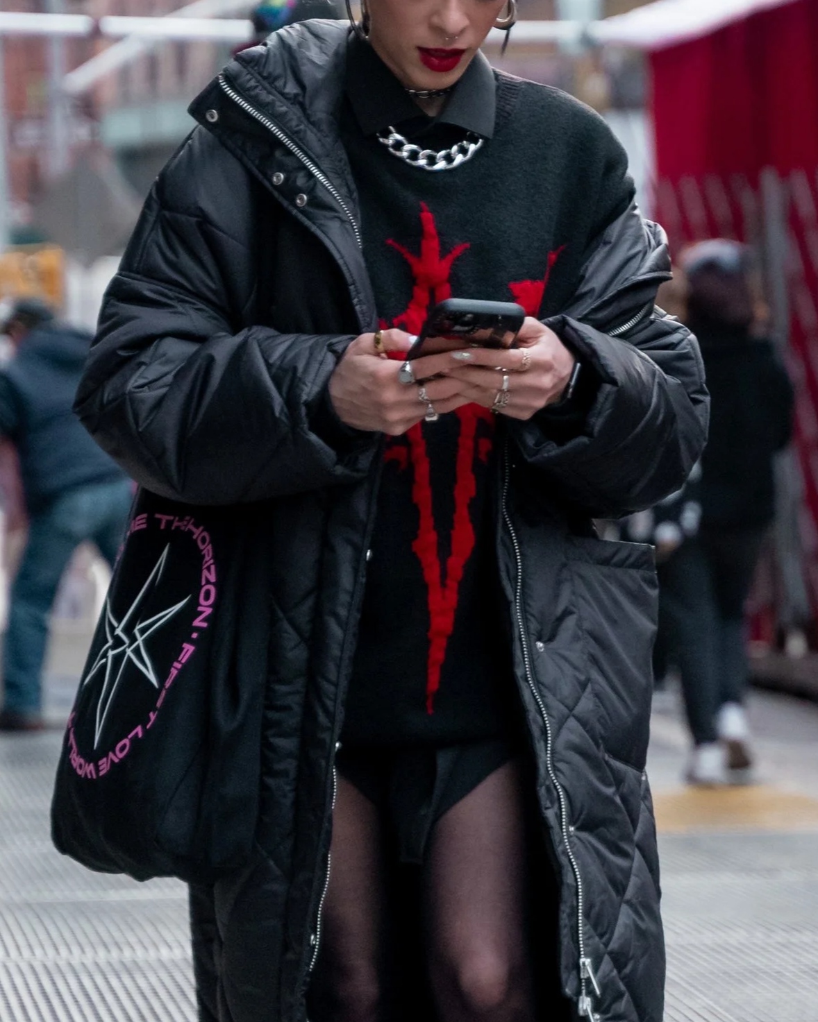 Person in black jacket and black graphic sweater with red design, wearing rings and a silver chain necklace, standing outdoors and using a smartphone.