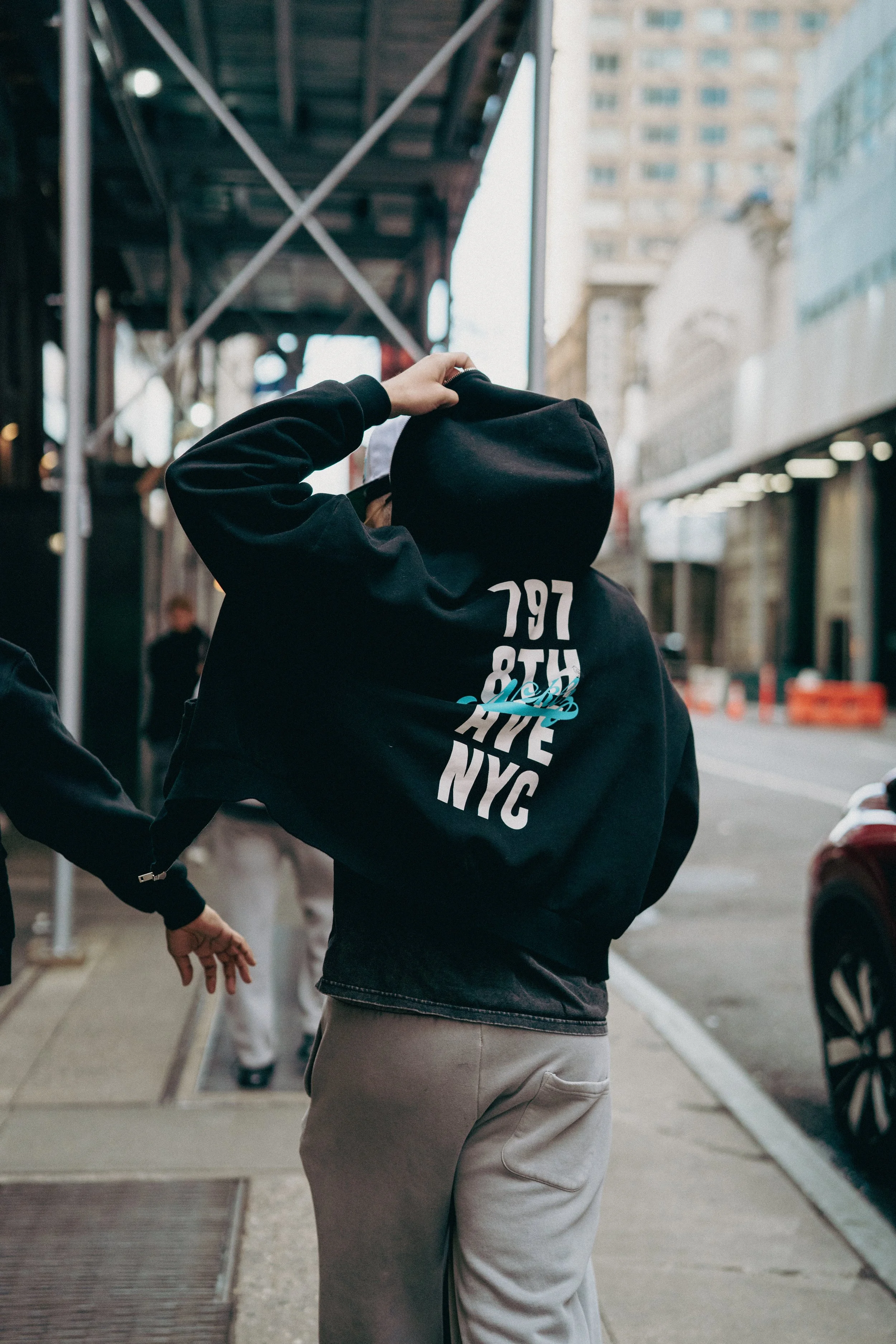 Person wearing a black hoodie with "797 8TH AVE NYC" printed on the back, standing on a city sidewalk with buildings and cars nearby.