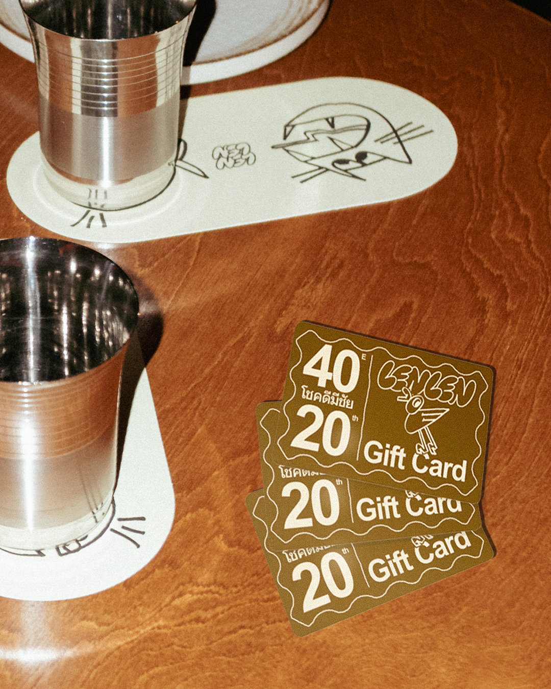 Three brown gift cards with the text '40,' '20,' and '20,' and the words 'Gift Card,' placed on a wooden table next to two metal cups. The table also has a paper with a cartoon drawing of a cat's face.