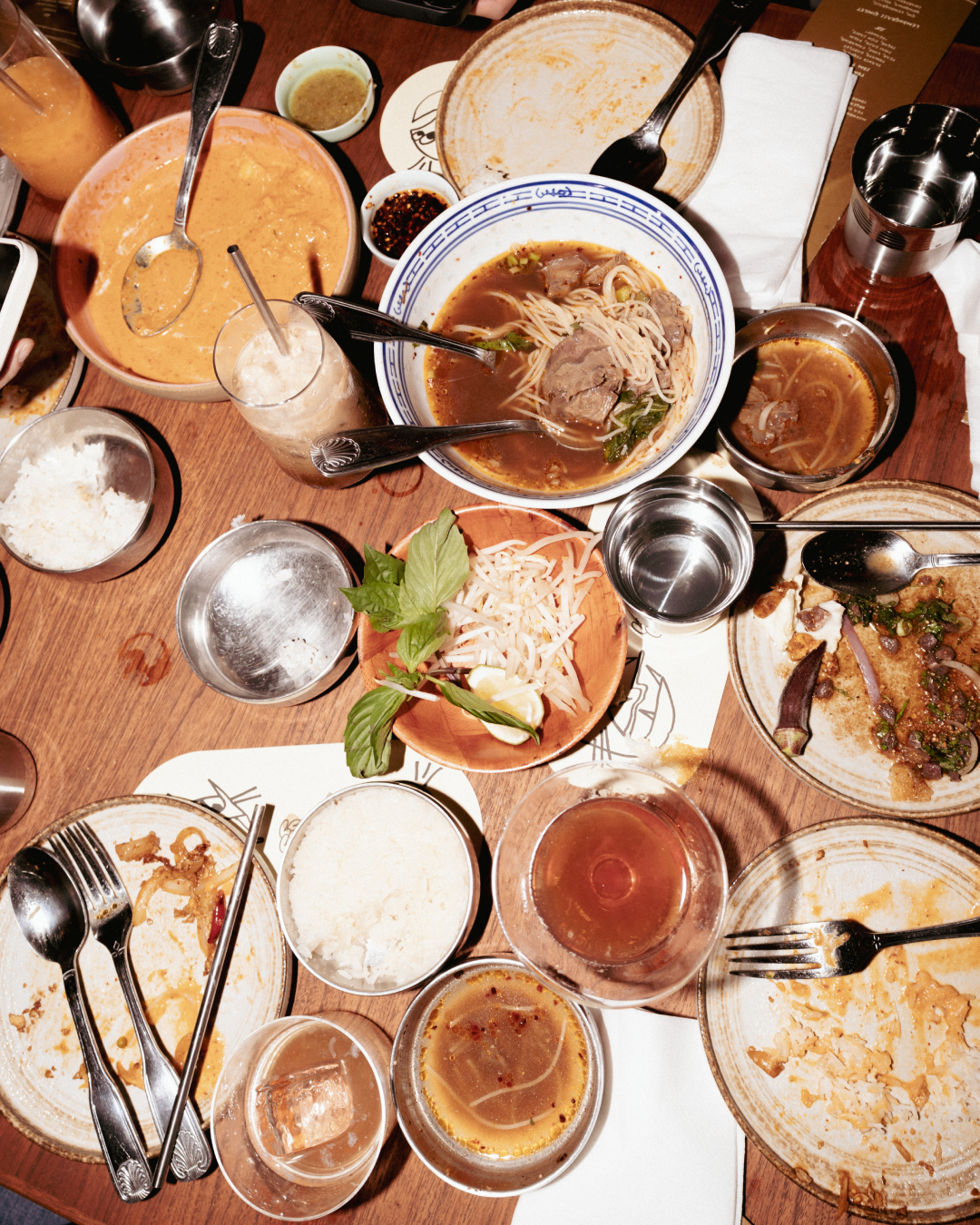 Empty and partially eaten plates of Asian cuisine, including noodle soup, rice, and various side dishes, on a wooden table.