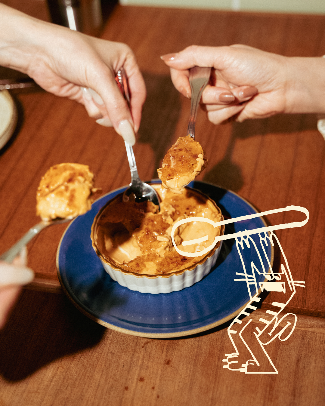 People serving themselves from a ramekin of dessert, with a spoon and fork, on a wooden table.