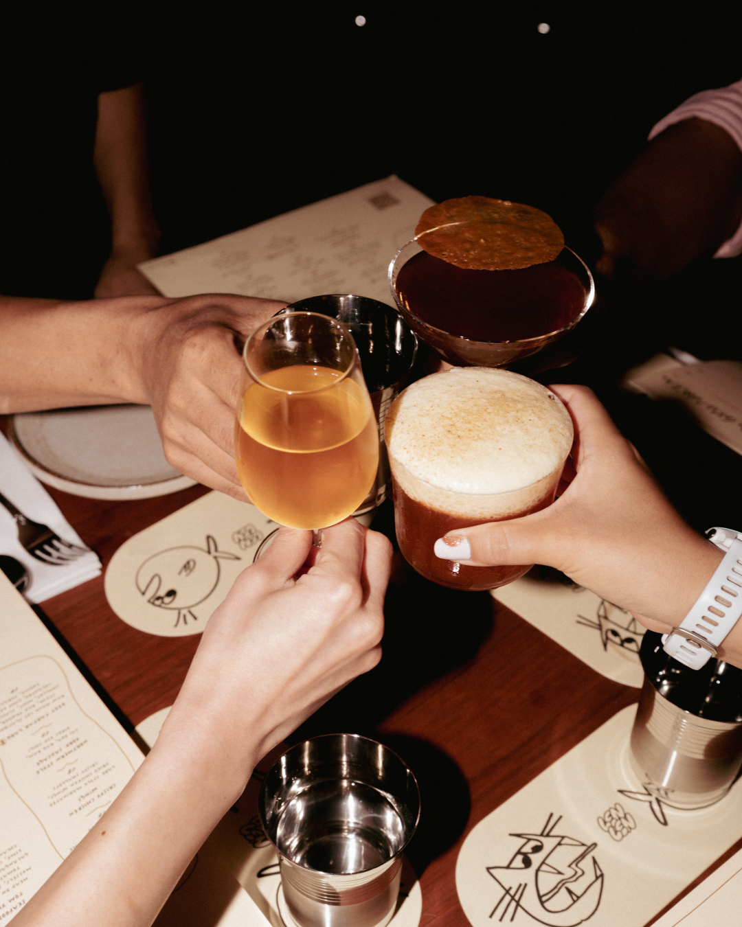 People holding various drinks in a toast at a restaurant table with menus and illustrated placemats.