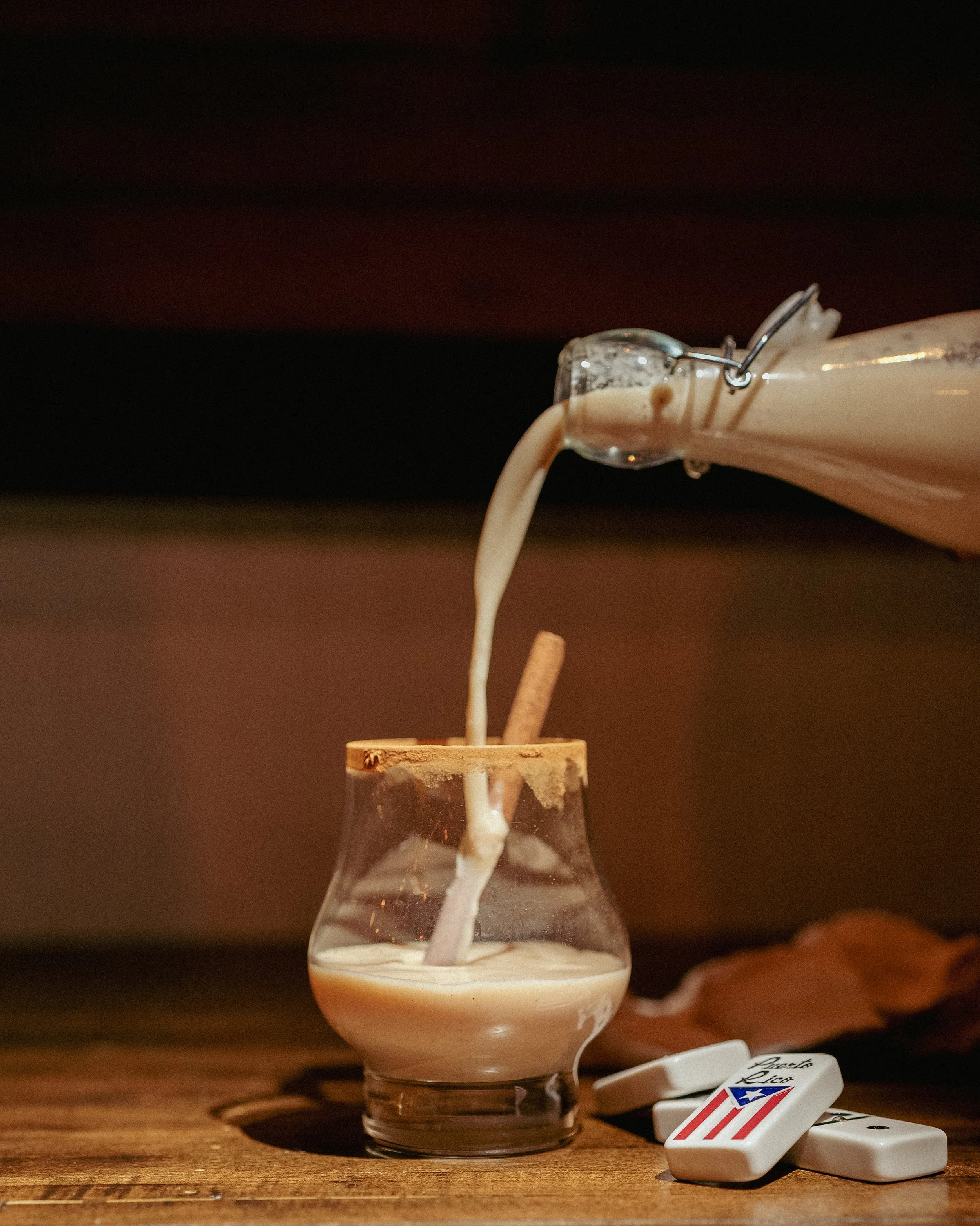 A glass of coffee with a cinnamon stick being filled with cream from a teapot.