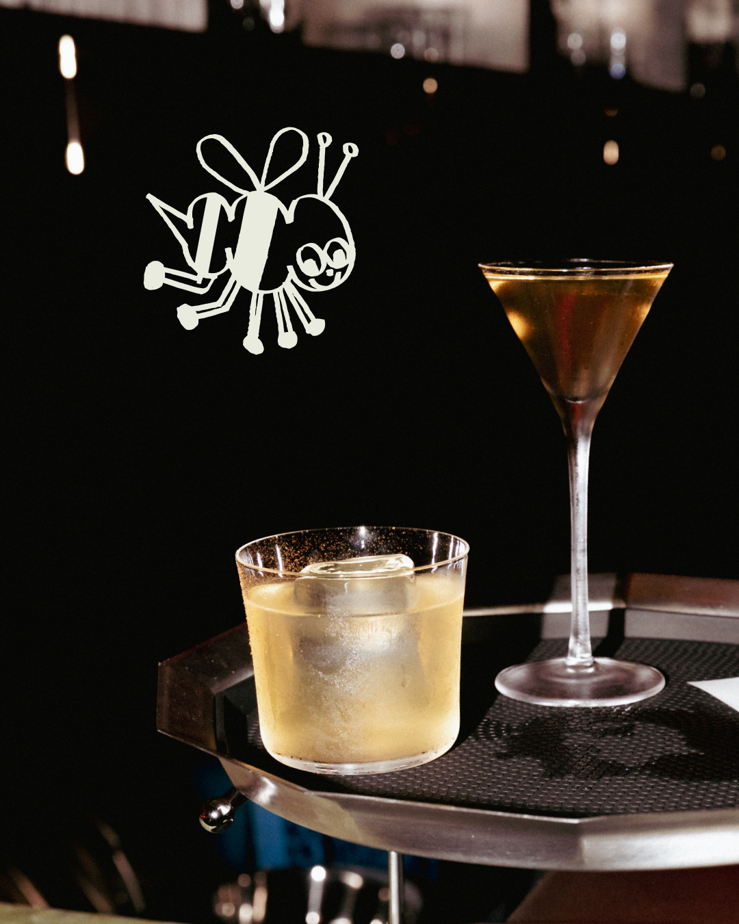 A hot foam drink with ice cubes in a glass and a dark cocktail in a martini glass on a black serving tray, with a cartoon bee illustration floating above.