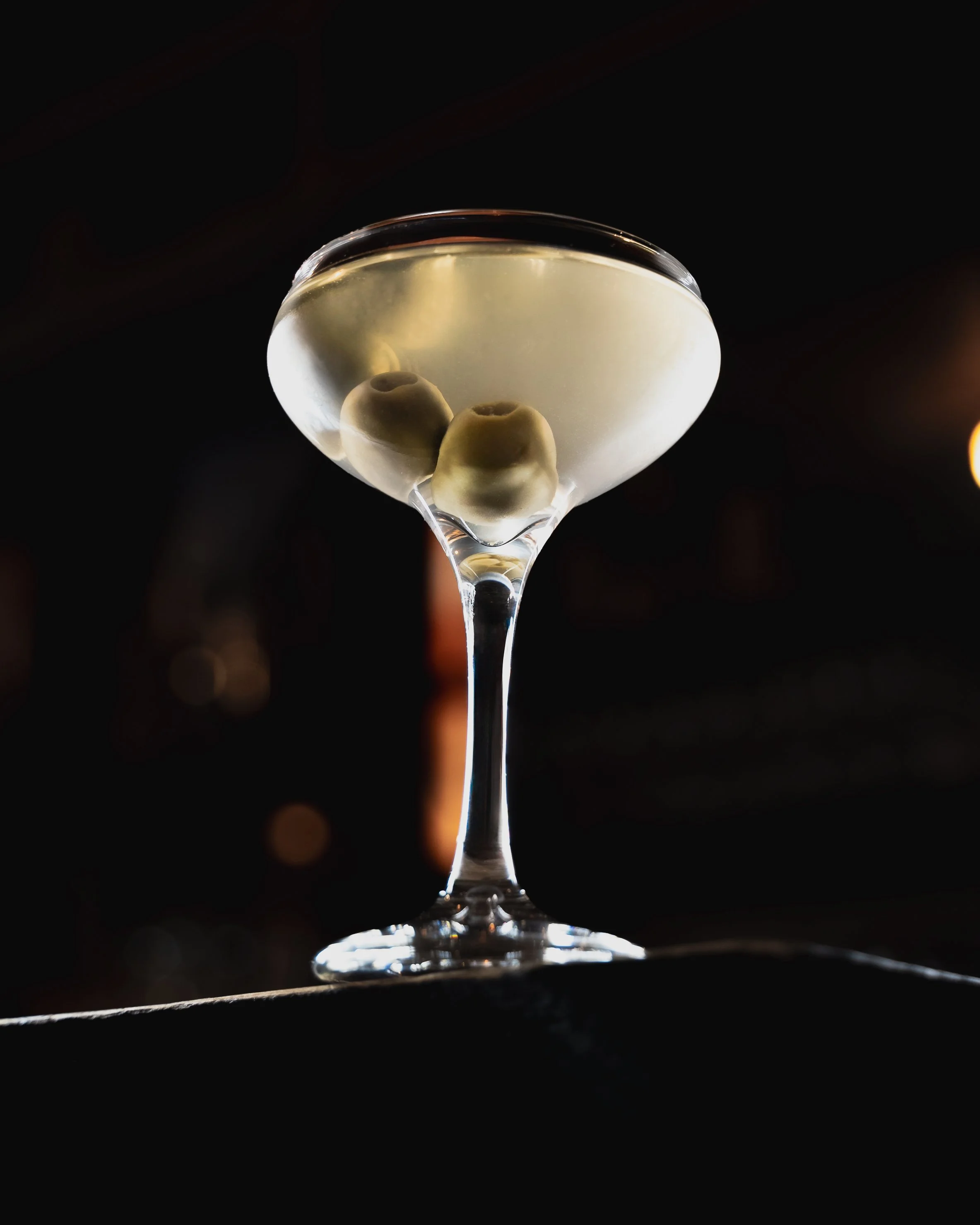 A cocktail glass with olives submerged in a clear liquid, set against a dark background.