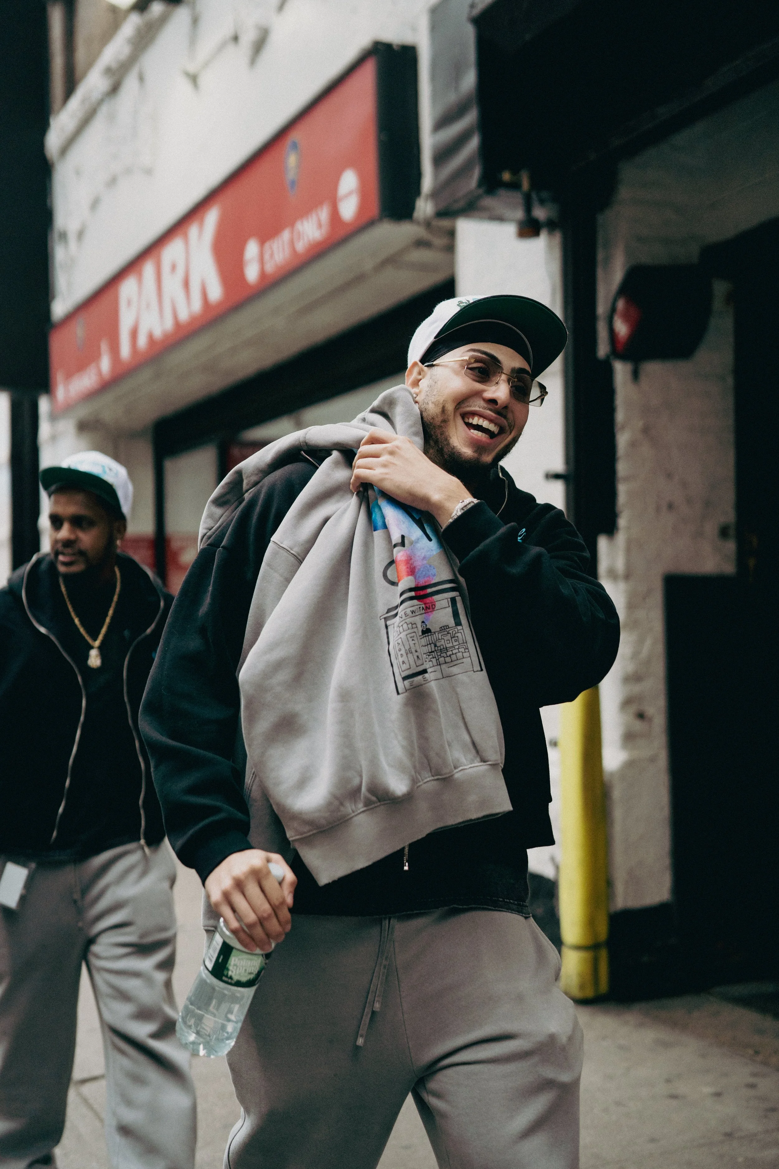 Two young men walking outside the entrance of a store with a red sign that says 'PARK'. The man in the foreground is smiling, wearing glasses, a black jacket, gray pants, and a gray hoodie, carrying a water bottle, and holding a beige hoodie on his s