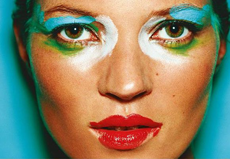 Close-up of a person's face with colorful, artistic makeup including blue eyeshadow, white accents, green lines, and red lipstick.