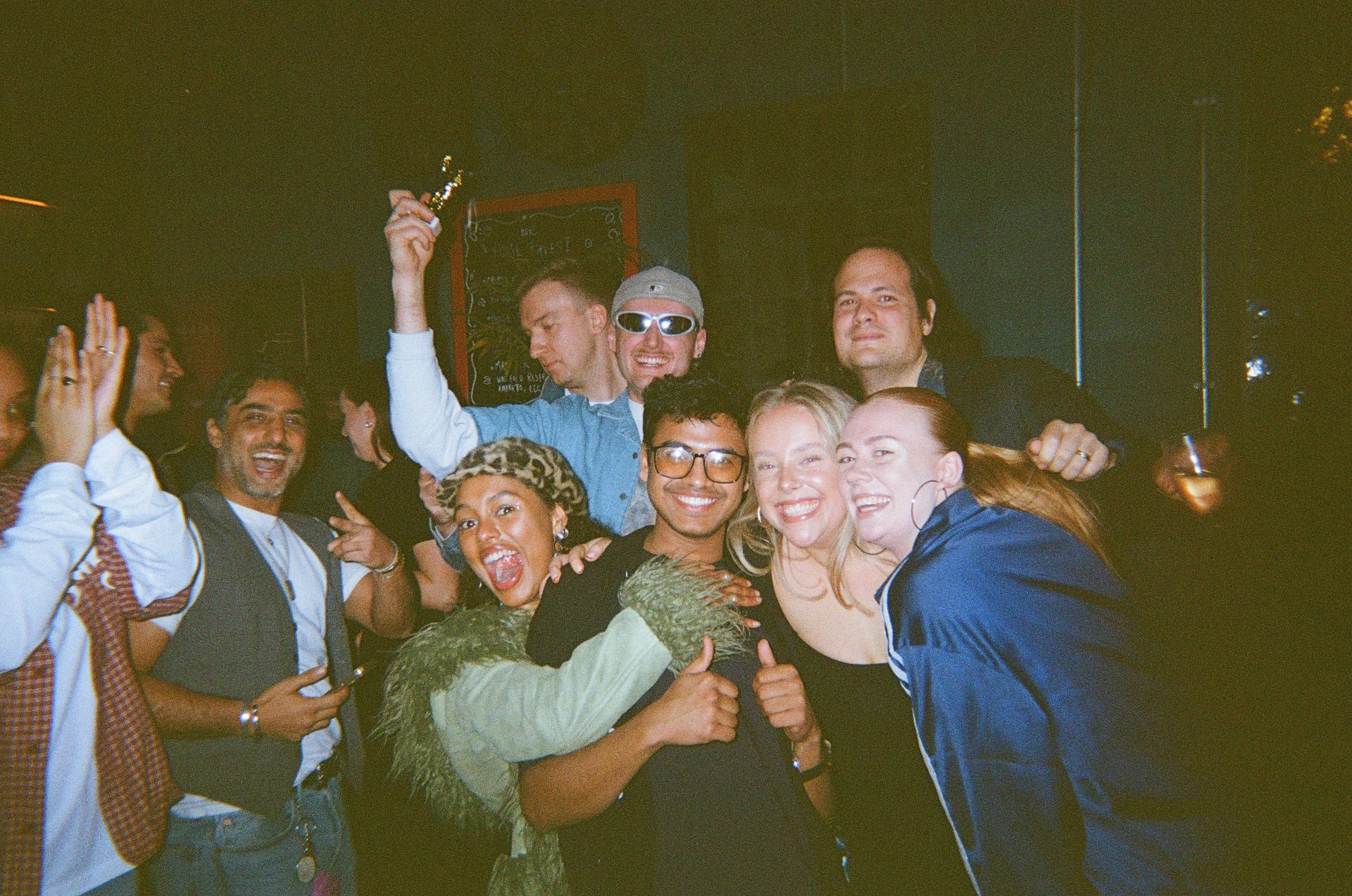 A diverse group of people smiling and celebrating together at a party or event, with some raising their hands and showing excitement.