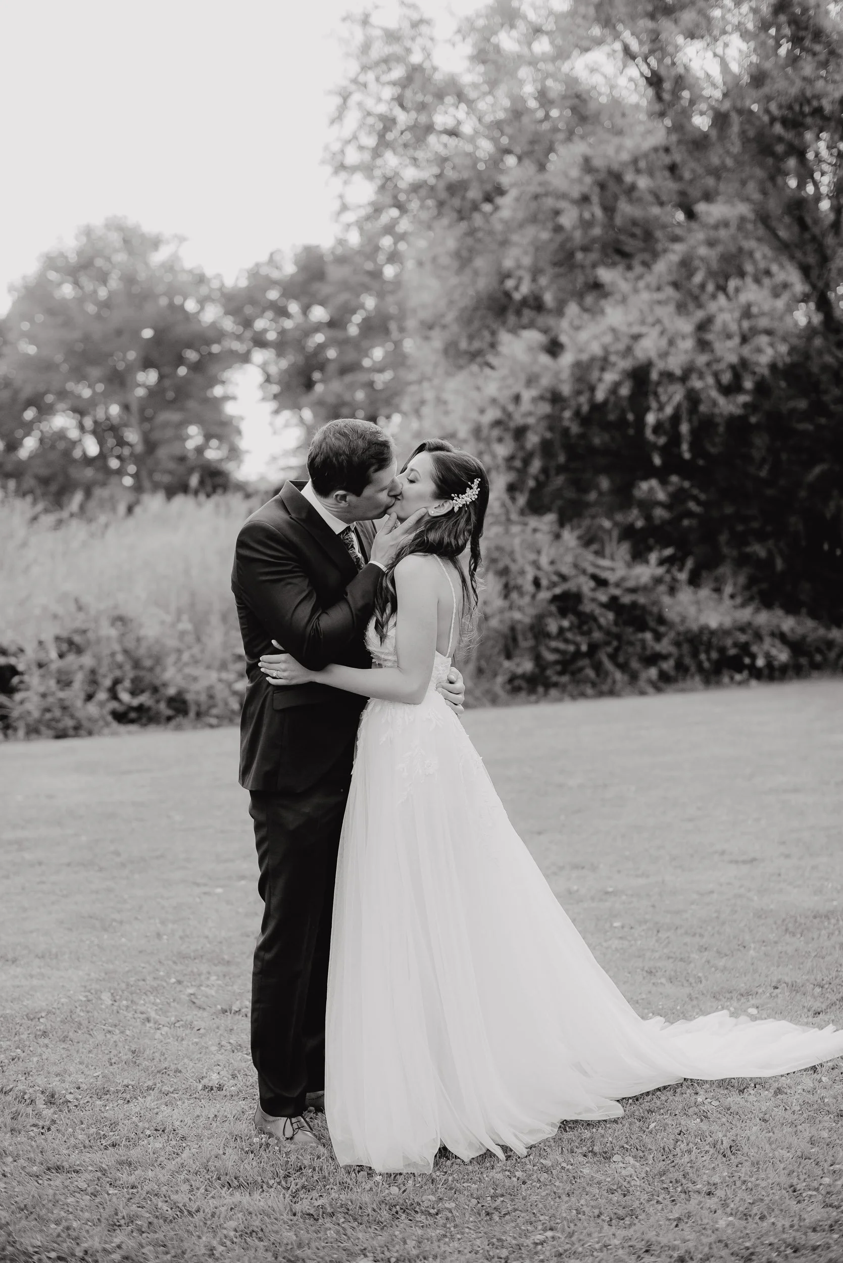 A black-and-white photo of a man and woman in wedding attire sharing a kiss outdoors. The man is in a suit and the woman in a wedding dress with a veil and floral hair accessory, standing on a grassy area with trees in the background.