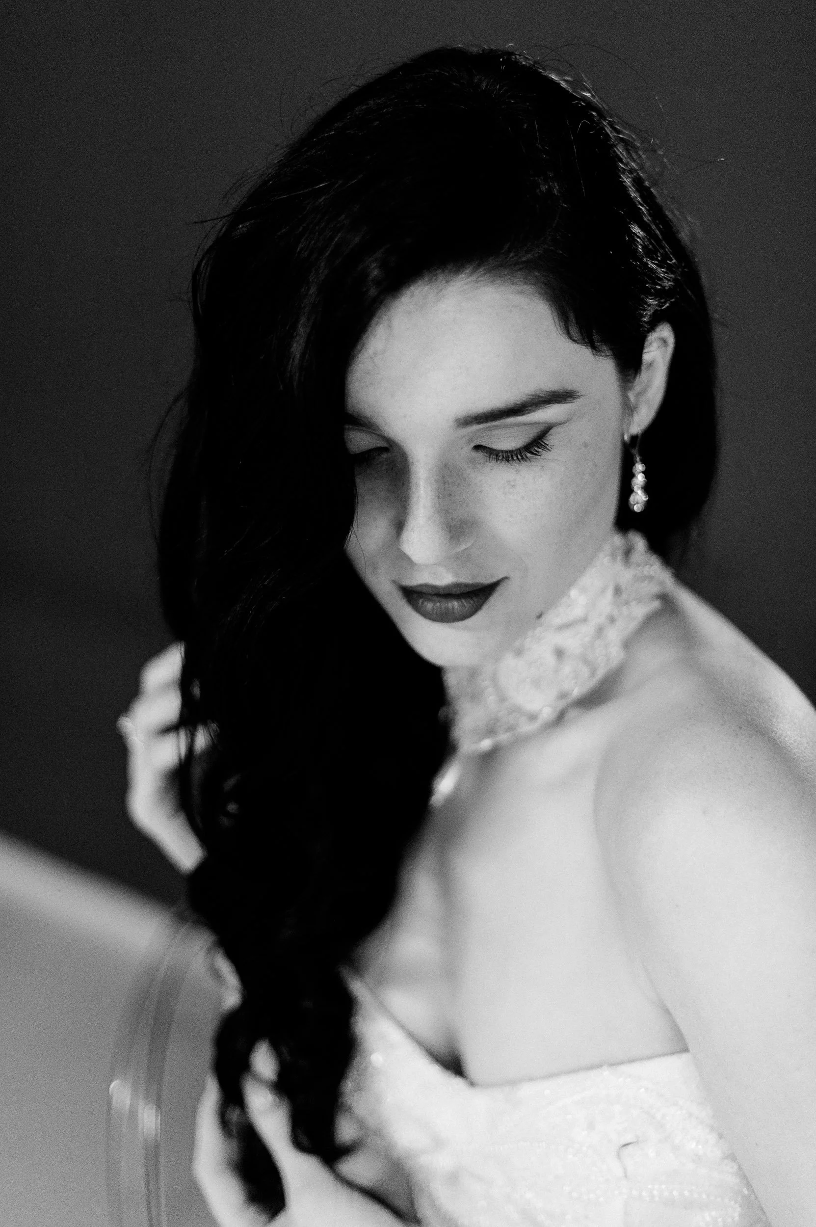 Black and white portrait of a woman with long, dark hair styled in loose waves, wearing earrings and a lacy dress, with her eyes closed and head slightly tilted down.