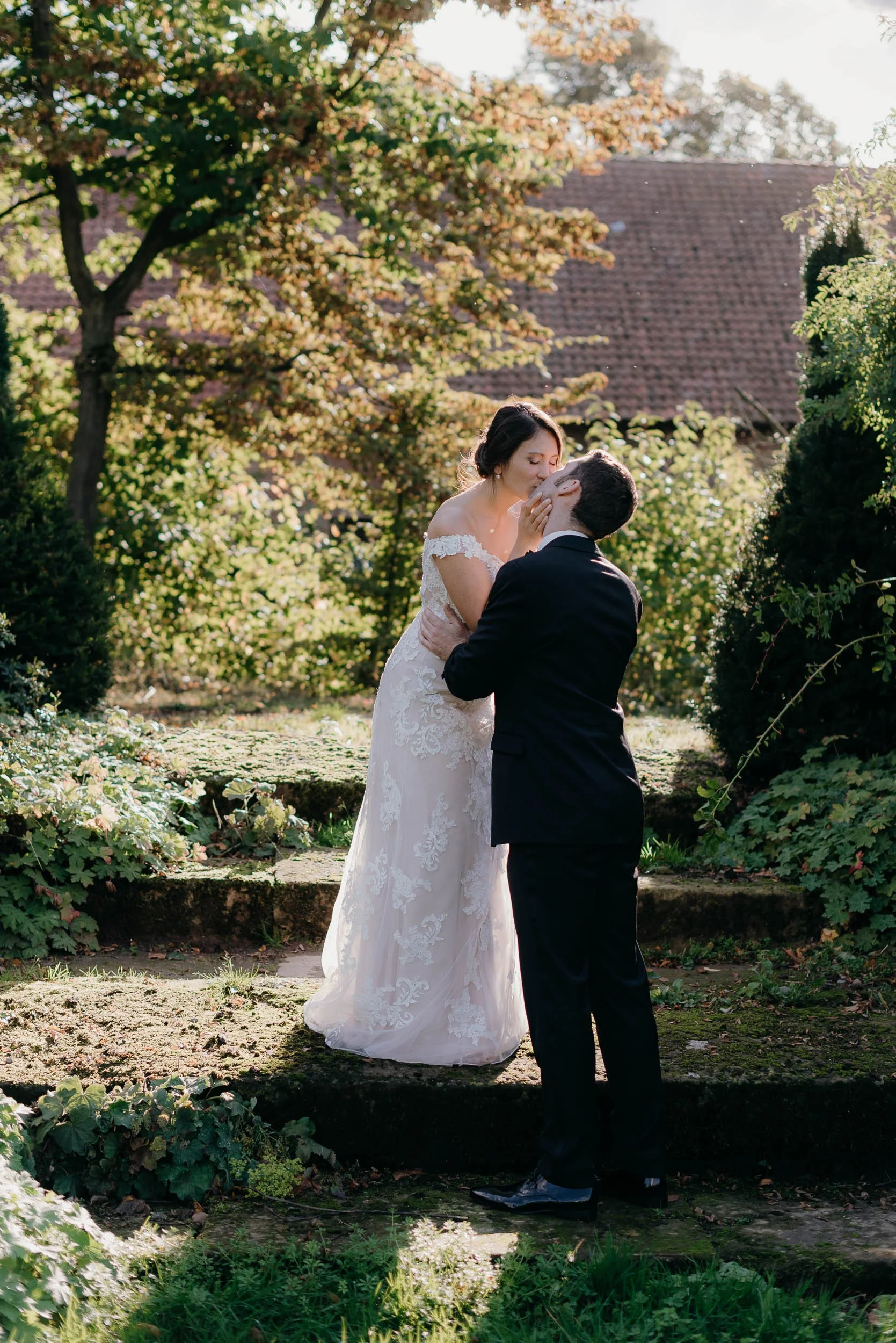 A bride and groom sharing a kiss outdoors in a garden with lush greenery and trees in the background during daylight.