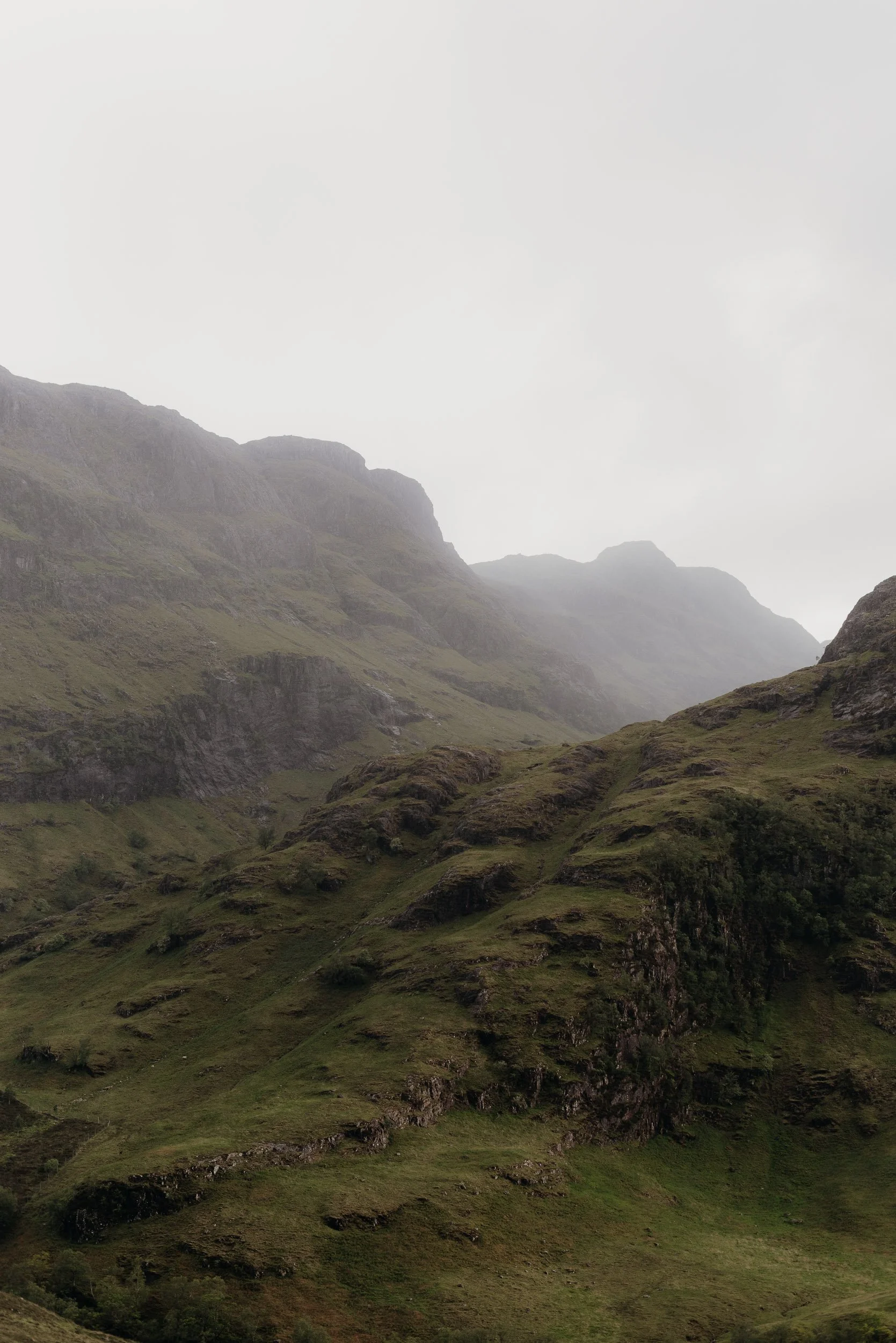 A landscape of green mountain hills with some rocky formations, shrouded in fog or low clouds under a pale, overcast sky.