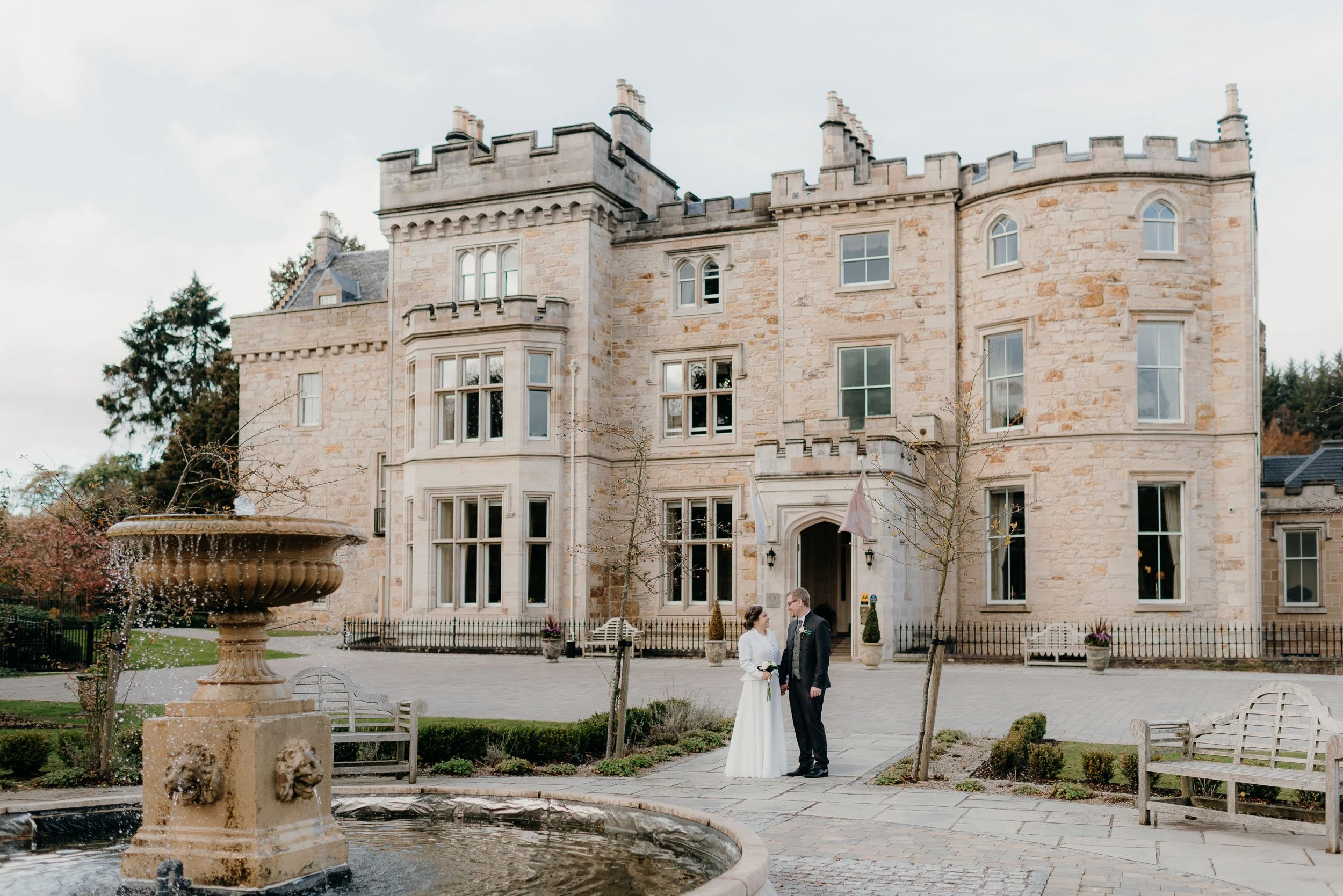 A bride and groom standing in front of a large stone castle-like building, with a fountain in the foreground and trees around.