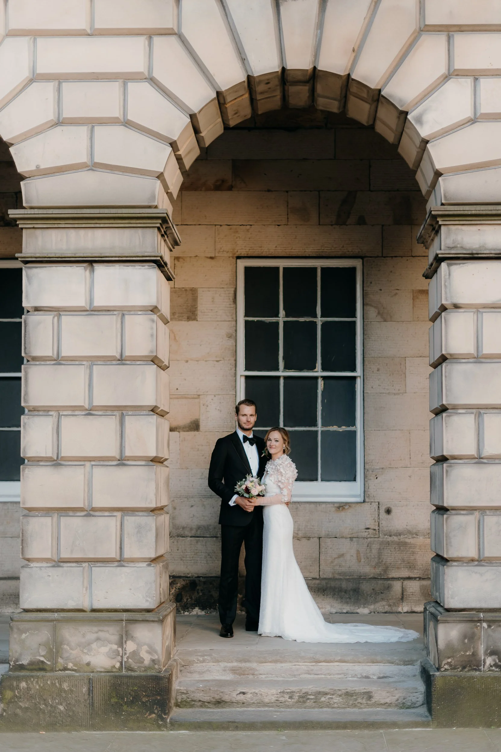 A wedding couple standing under a stone archway in front of a large window, with the groom in a black tuxedo and the bride in a white wedding dress holding a bouquet.