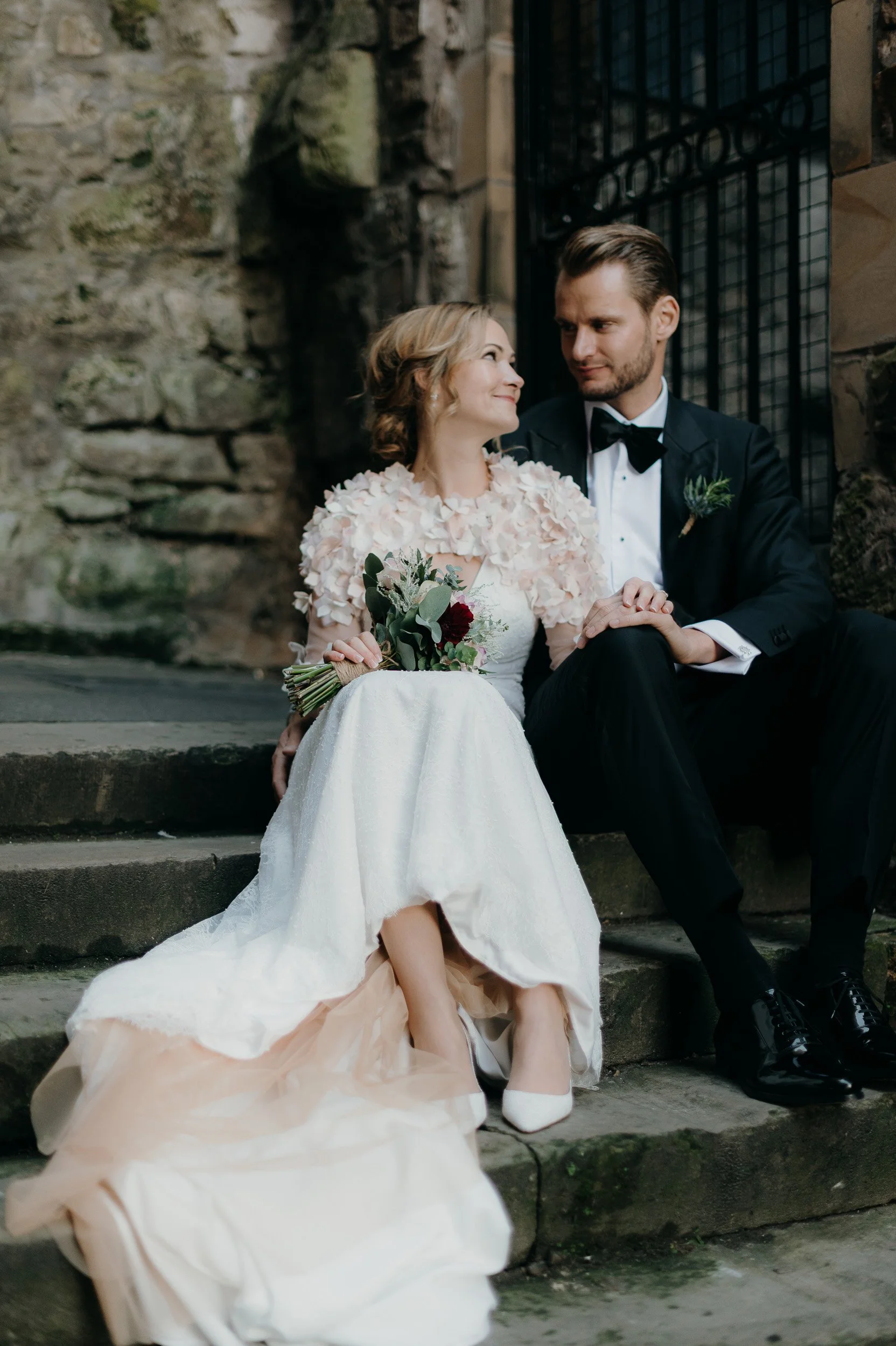 A bride and groom sitting on stone steps, gazing into each other's eyes. The bride is wearing a white gown with floral details and holding a bouquet, while the groom is dressed in a black tuxedo with a bow tie. They are outdoors near a stone wall and black iron fence.
