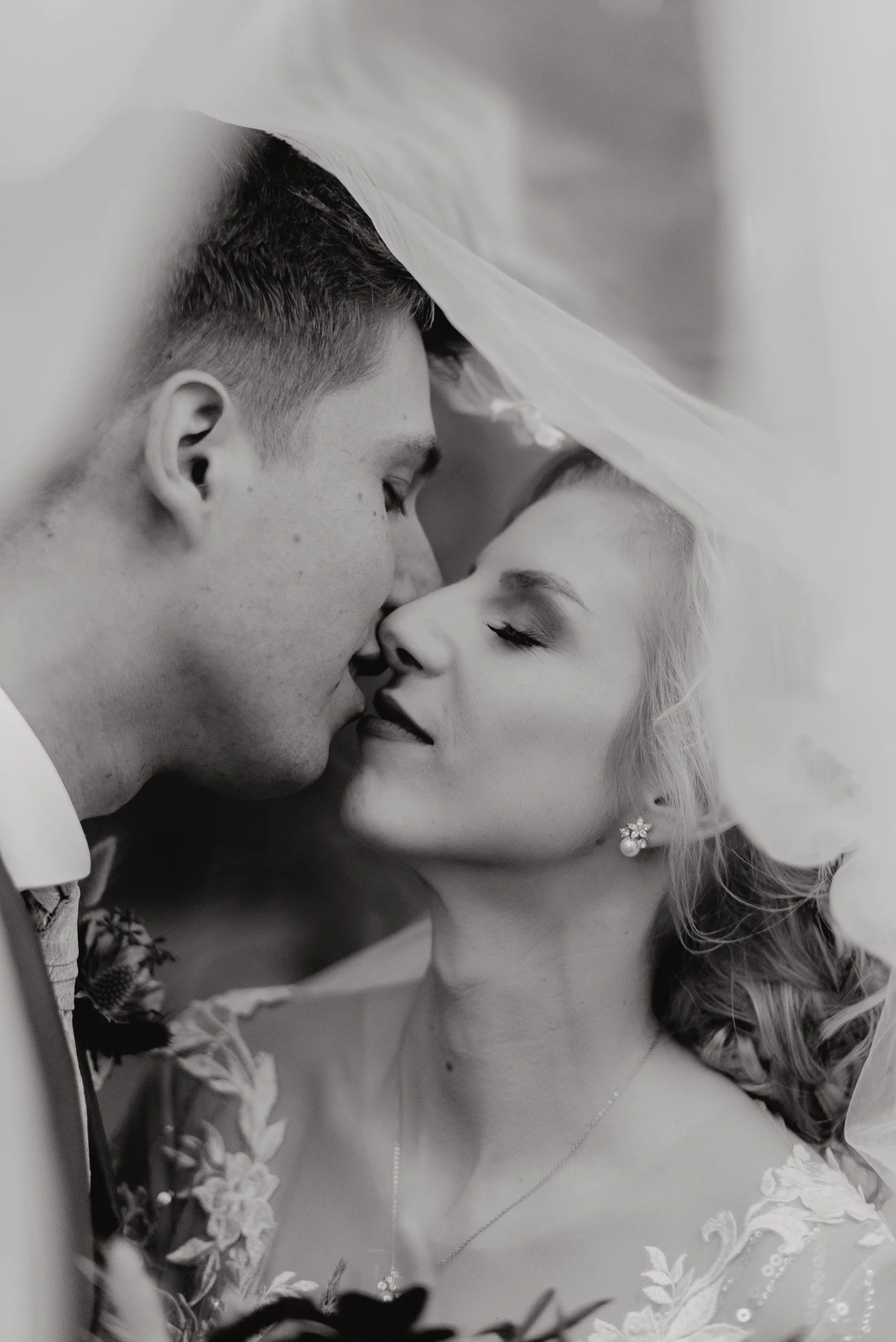 A black and white photo of a man and woman close to each other, about to kiss. The man has short hair and is dressed in formal attire, while the woman has long, curly hair, elegant earrings, and a floral dress. They are under a sheer fabric, possibly a wedding veil.