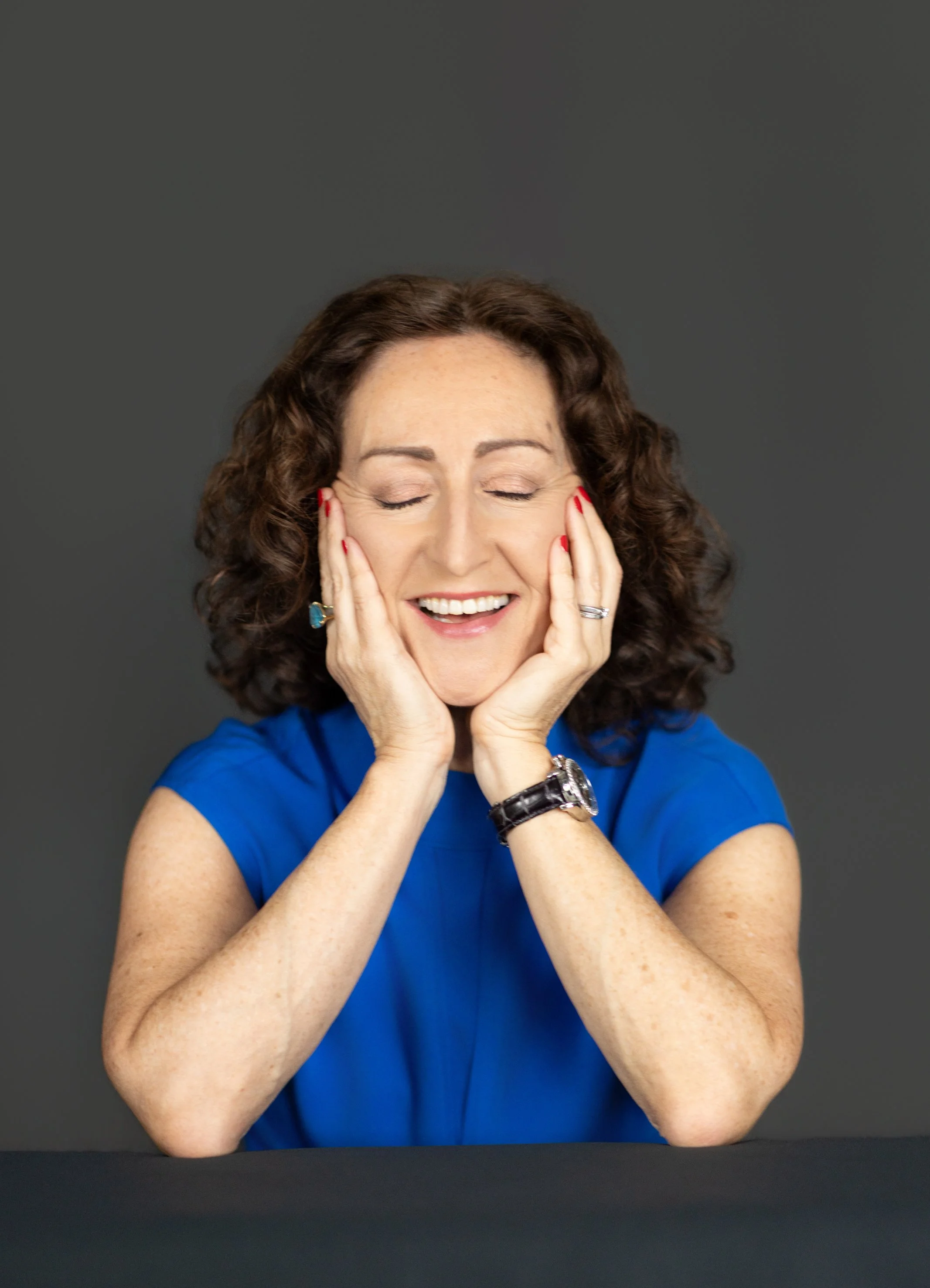 A woman with curly brown hair, wearing a blue top, smiling with eyes closed, holding her face with both hands, against a dark gray background.
