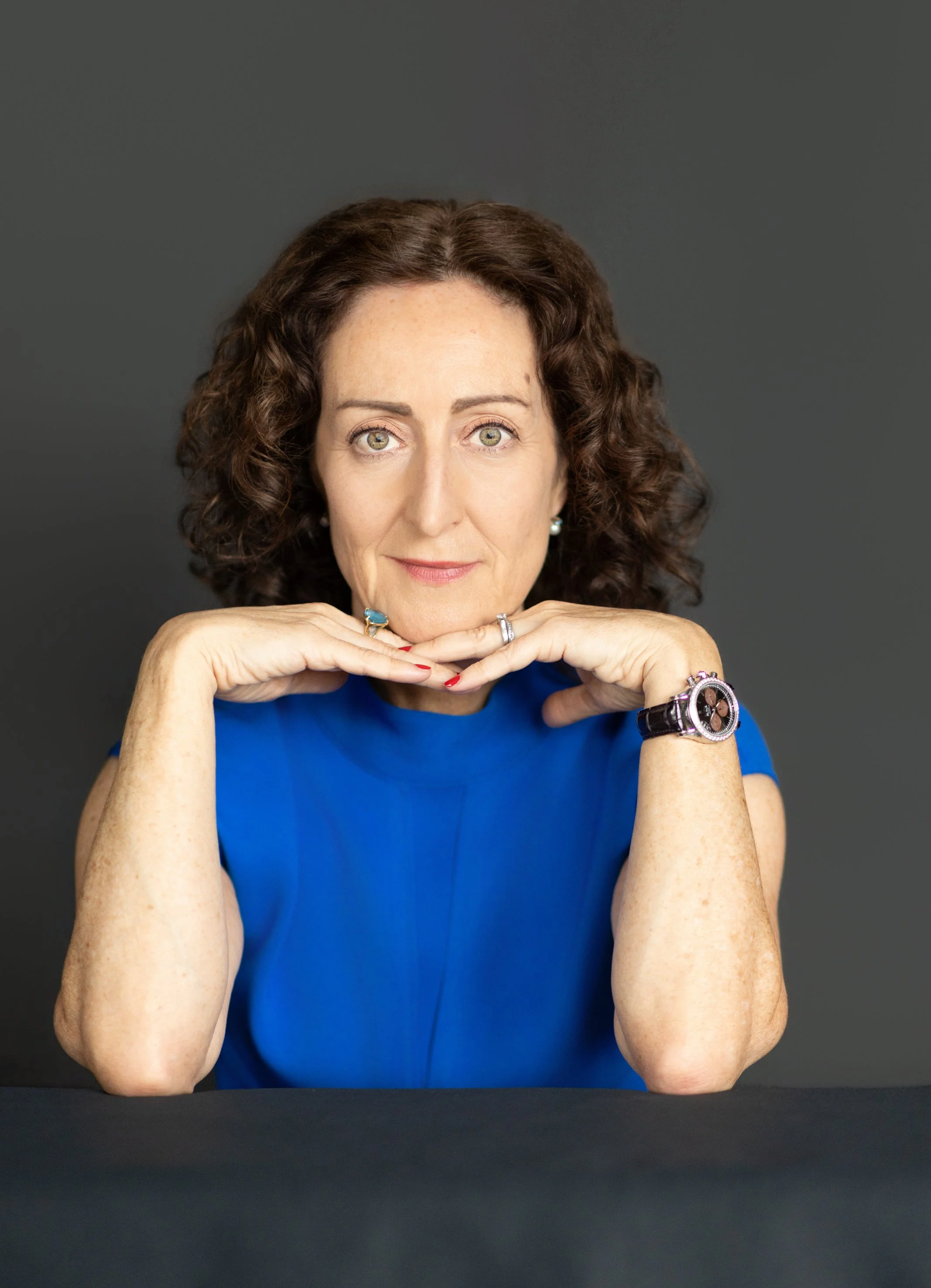 A woman with curly brown hair and light green eyes, wearing a blue top, resting her chin on her hands with rings on her fingers, wearing a watch, posing against a plain dark background.