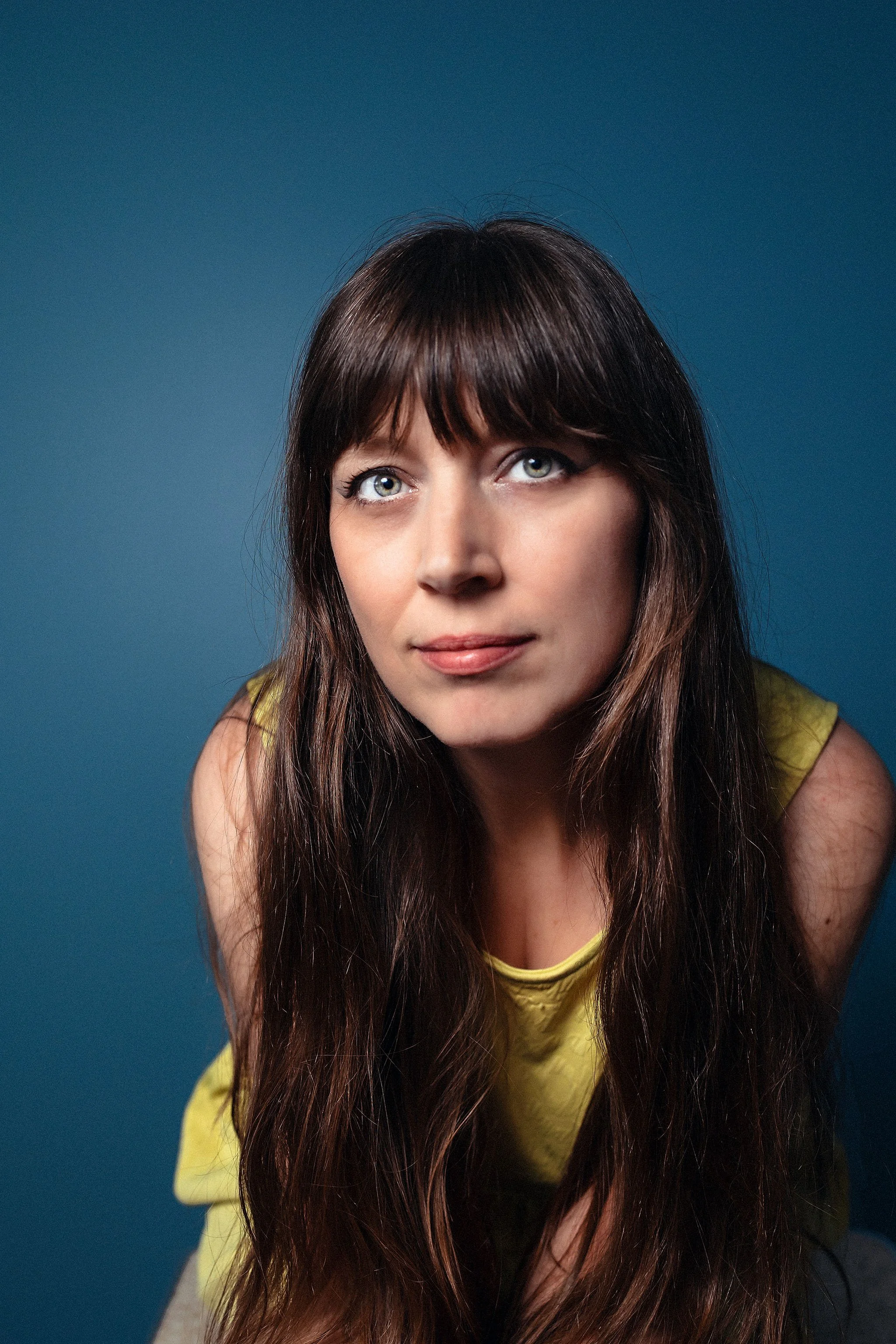 Close-up self-portrait of Heather Bays in a yellow top against a dark blue background, looking upward in soft directional light.