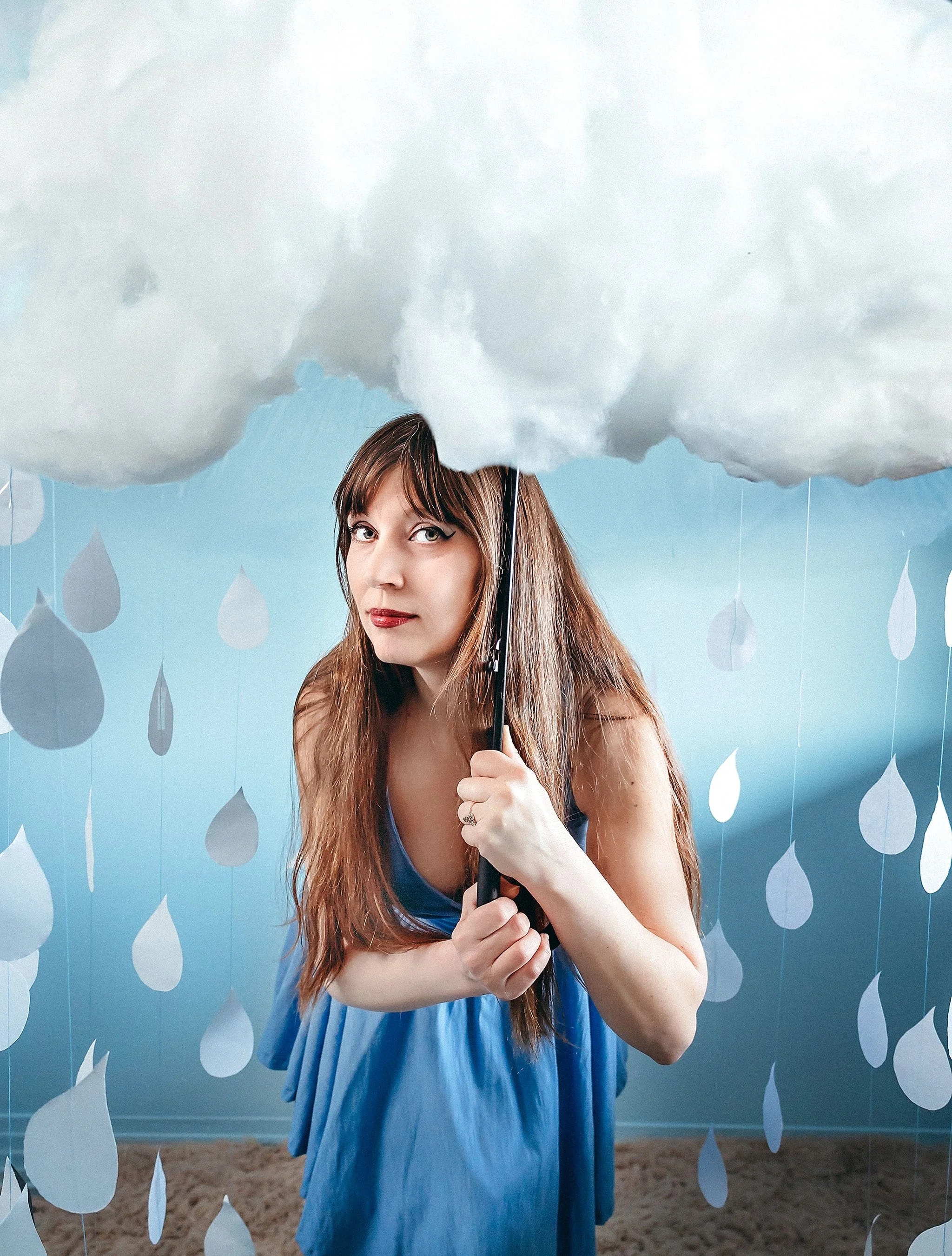 Conceptual self-portrait of Heather Bays in a blue dress holding a handmade cloud umbrella, surrounded by paper raindrops against a blue background.