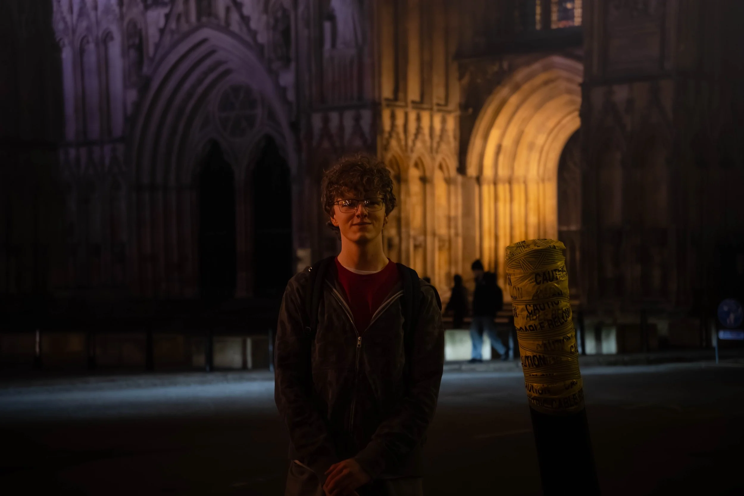 A young man with curly hair and glasses, standing outdoors at night in front of a large illuminated building with gothic architecture. There is a yellow caution tape wrapped around a pole nearby, and a few people in the background near the building.