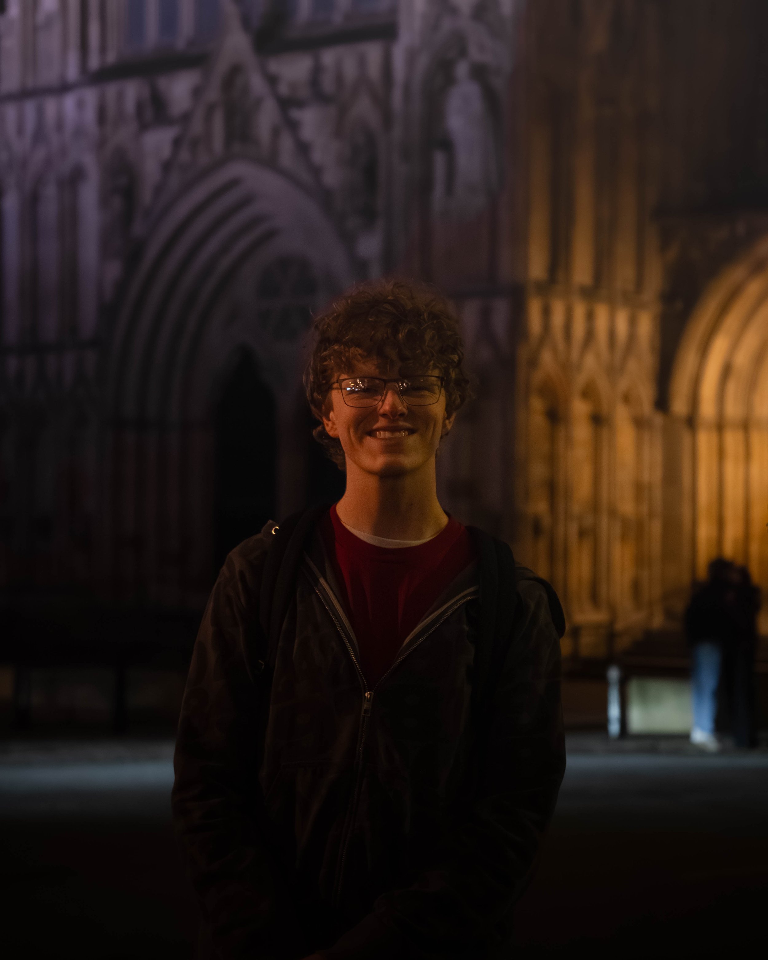 Young man with glasses smiling at night in front of a lit historic building with intricate stone architecture.