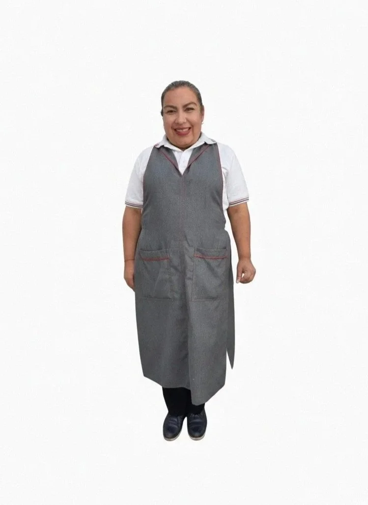 Mujer sonriendo con uniforme gris y camisa blanca, de pie frente a fondo blanco.