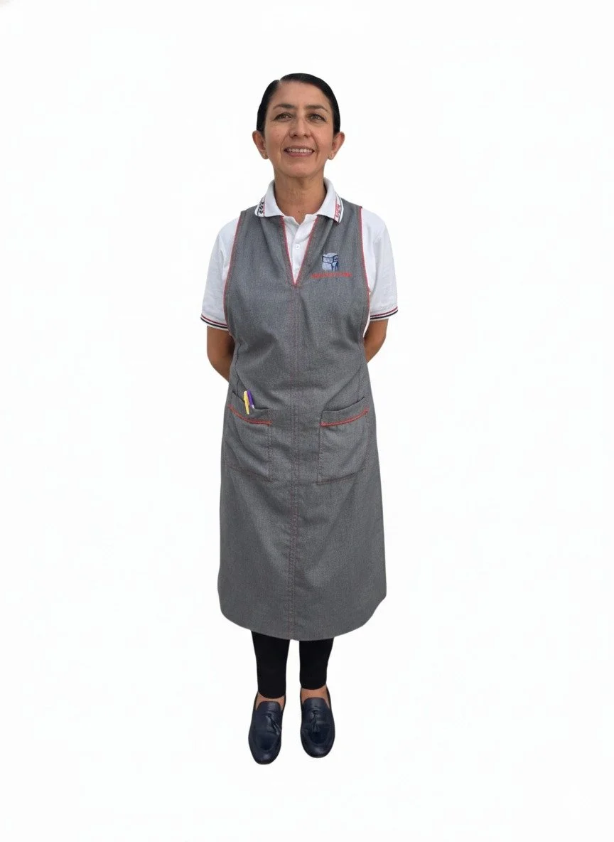 Mujer con delantal gris, camisa blanca y zapatos oscuros, de pie frente a fondo blanco.