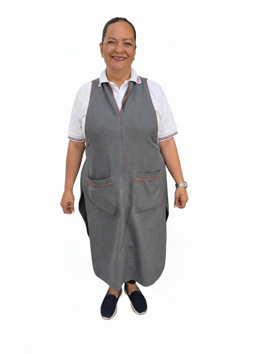 Mujer sonriendo, vestida con uniforme y un delantal gris, de pie contra fondo blanco.