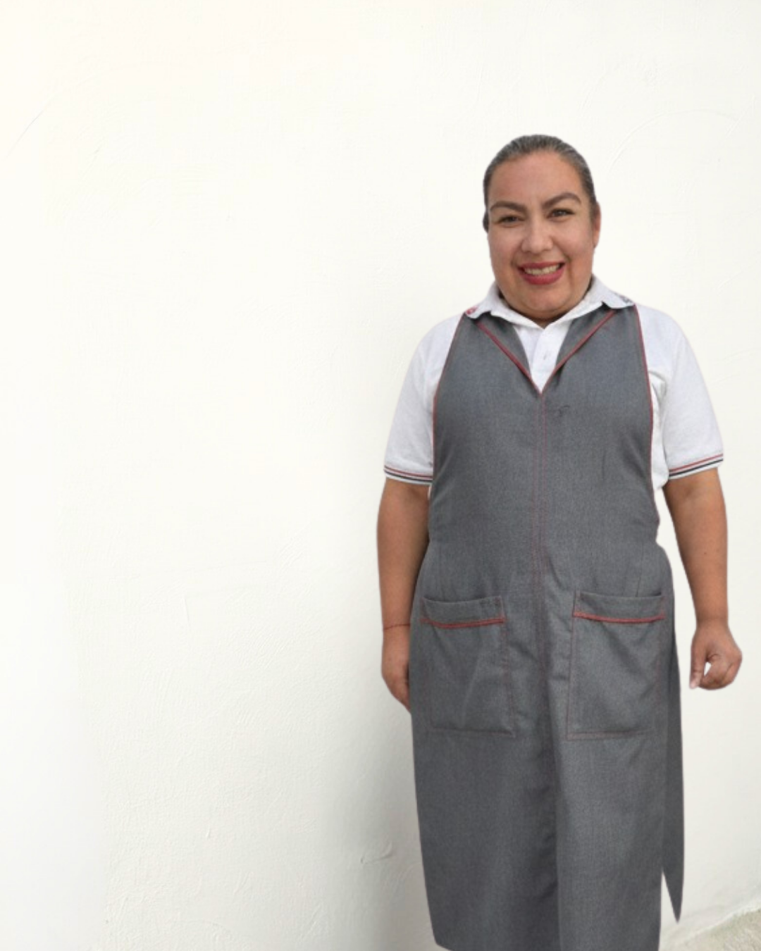 Mujer sonriendo con uniforme gris y camisa blanca, de pie frente a fondo blanco.