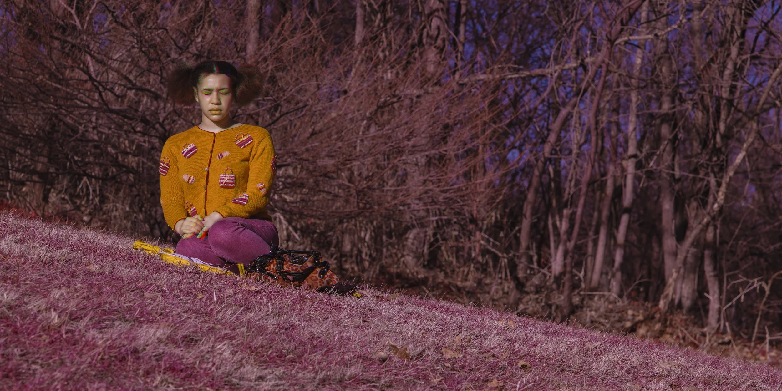 A young woman with brown hair styled in two pigtails, wearing a yellow cardigan with heart and candy patterns, sits cross-legged on a hill with pinkish grass. She has closed eyes, her hands clasped, and appears to be in a meditative pose, with a fore