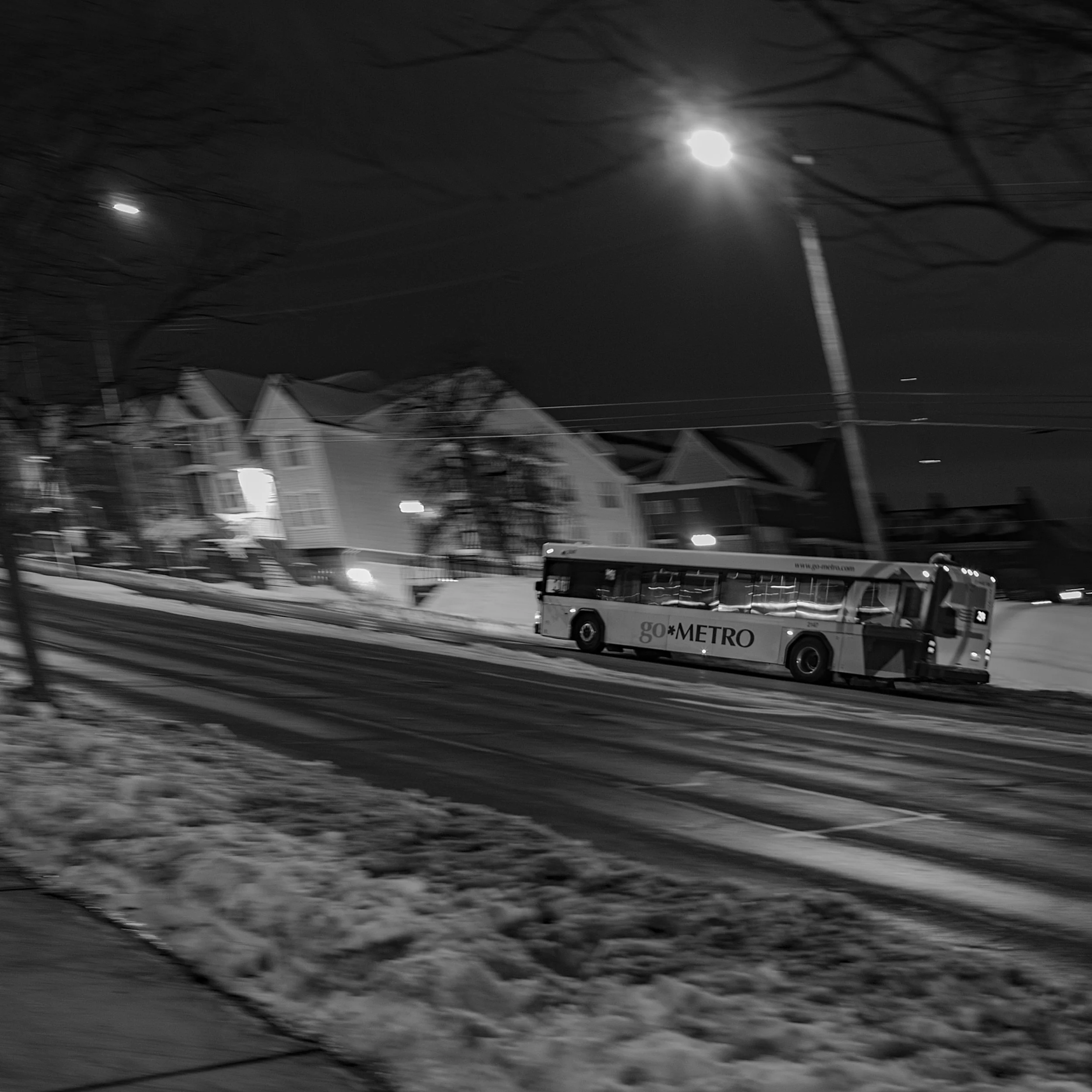 A metro bus driving on a wet, snowy street at night, with houses and powerlines in the background, illuminated by streetlights and a full moon.