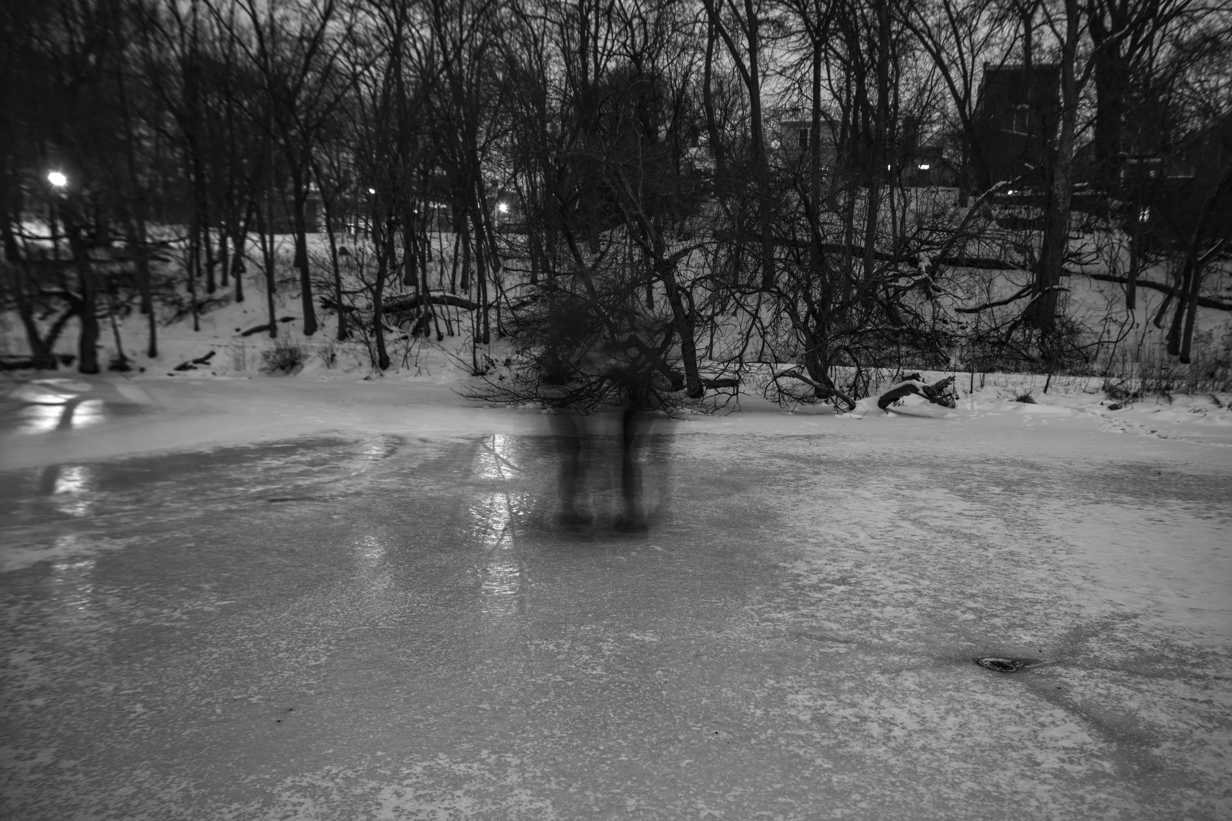 A blurred figure stands on a frozen, snowy pond with leafless trees and houses in the background, during nighttime.