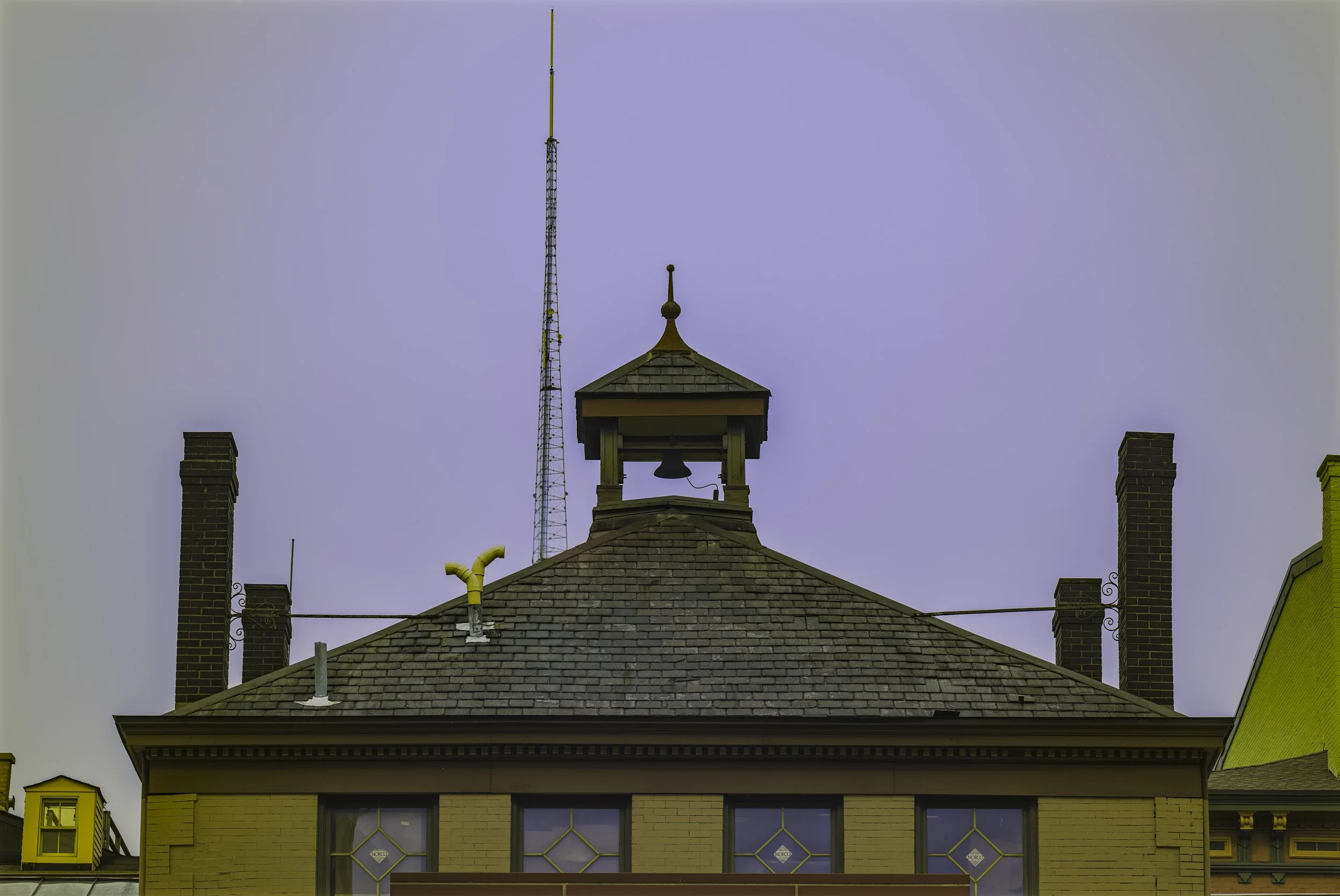 The roof of a yellow building with three windows and a small clock tower on top, topped with a weather vane and a lightning rod, with a tall antenna in the background and the sky above.