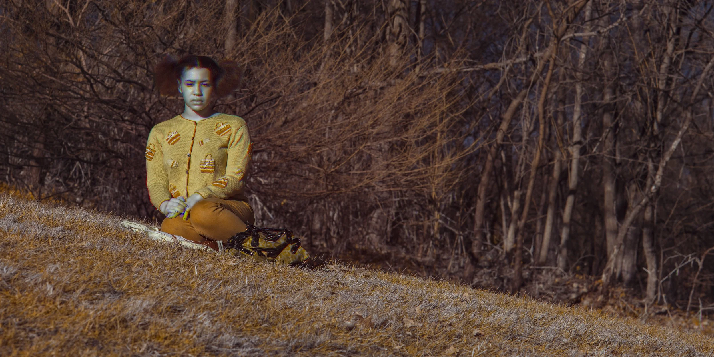 A young girl with puffy hair, wearing a yellow sweater with cupcake patterns, sitting on a grassy hill with a backpack beside her, in front of leafless trees during fall or winter.
