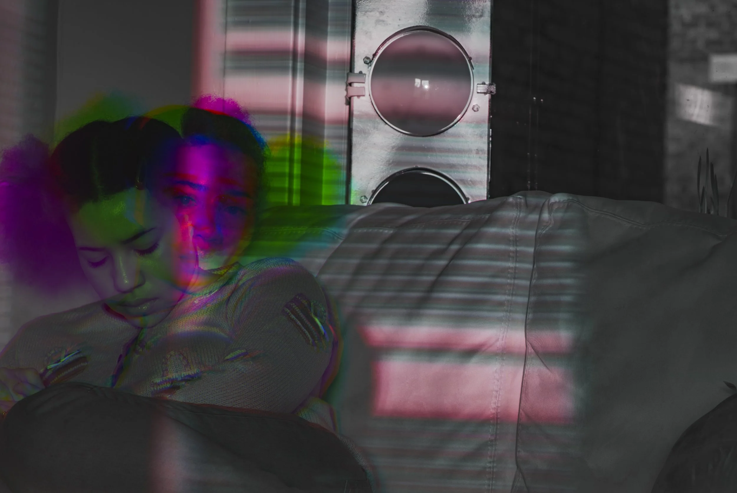 A black-and-white photo of a woman sitting on a sofa with multicolored light streaks and a reflection on her face, next to a metal speaker and a brick wall.