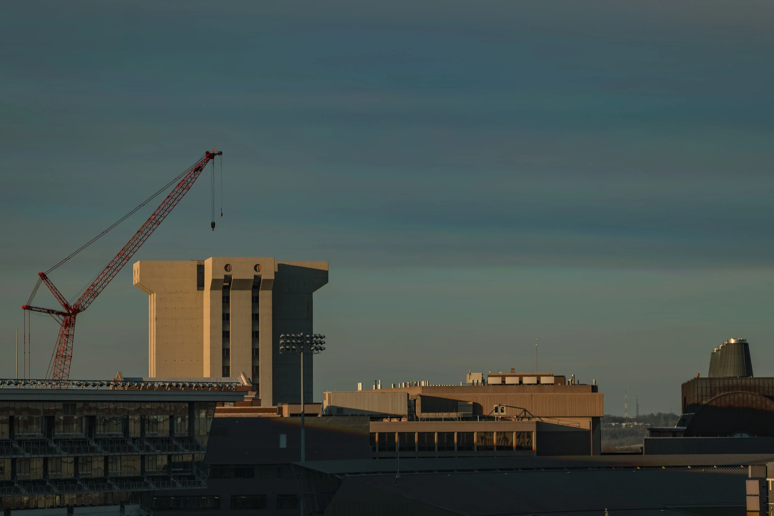 City skyline with a tall, unique concrete building, a construction crane, and other modern buildings under a blue sky with streaked clouds.