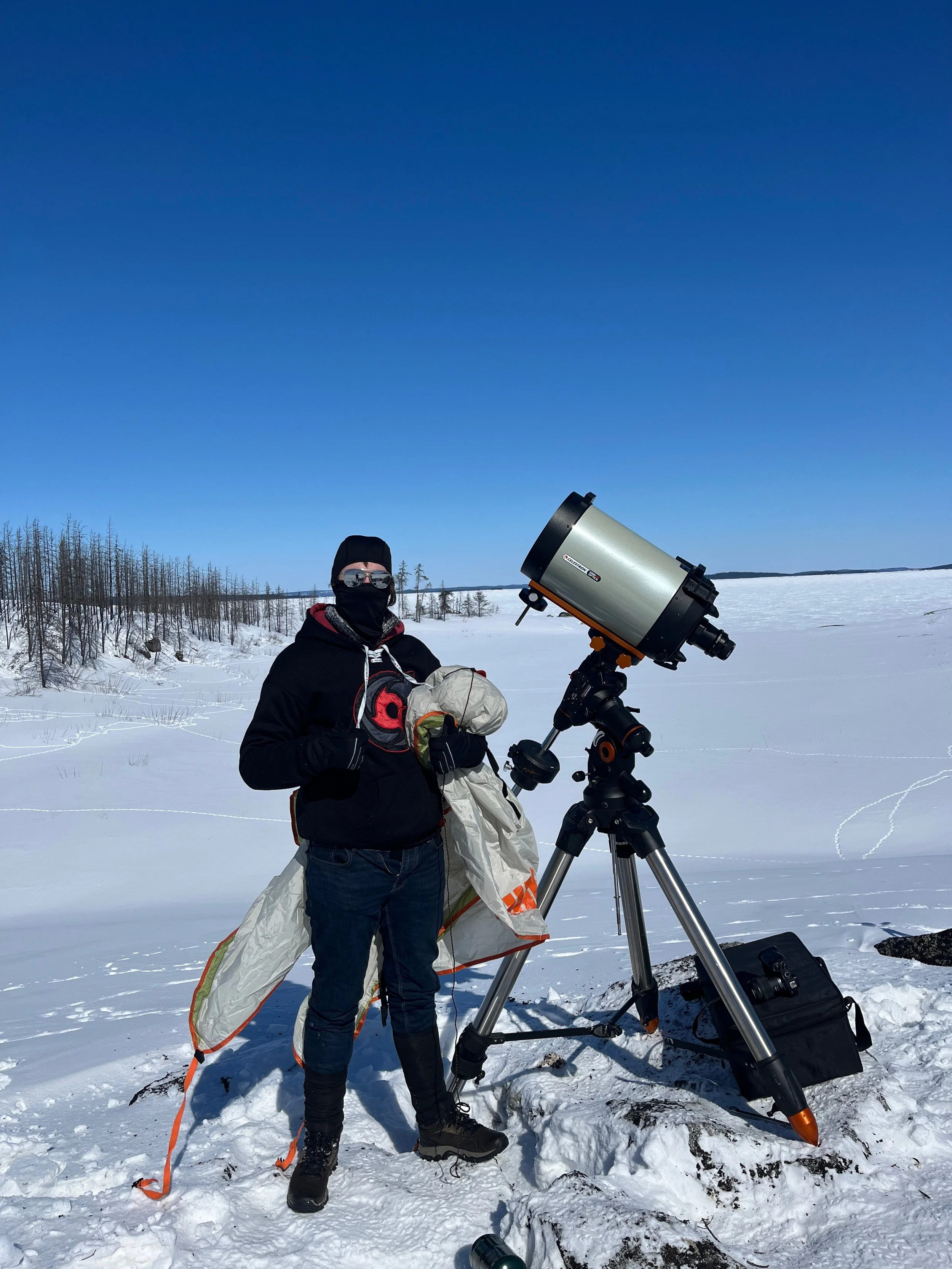 A person dressed in winter clothing and protective gear stands in a snowy landscape next to a large telescope on a tripod, holding a pack and standing near a black bag.