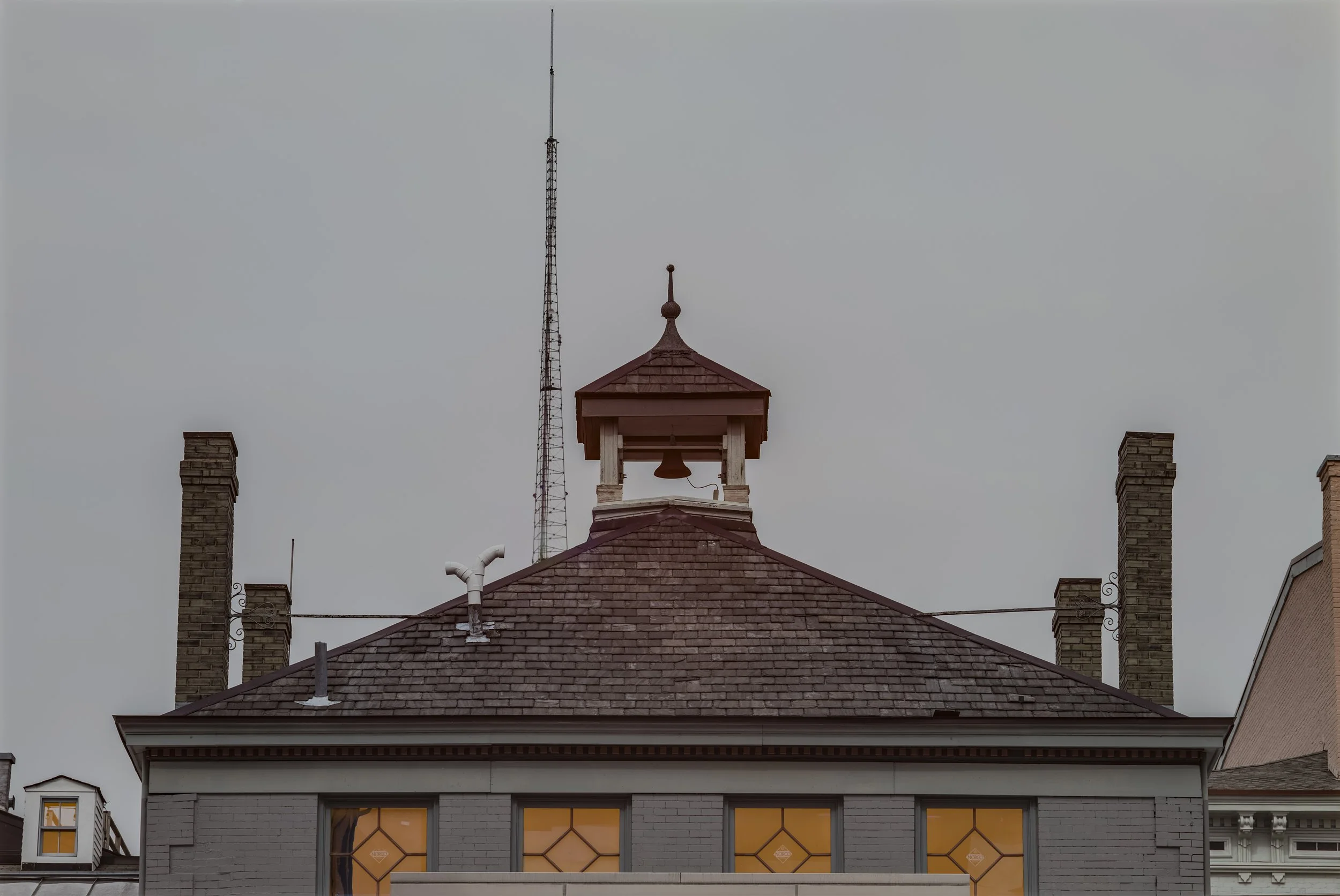 The image shows the rooftops of two older buildings with a cloudy sky in the background. The target building has a grey brick exterior, four windows with yellowish curtains, and a distinctive small bell tower on top of the roof. Two brick chimneys ar