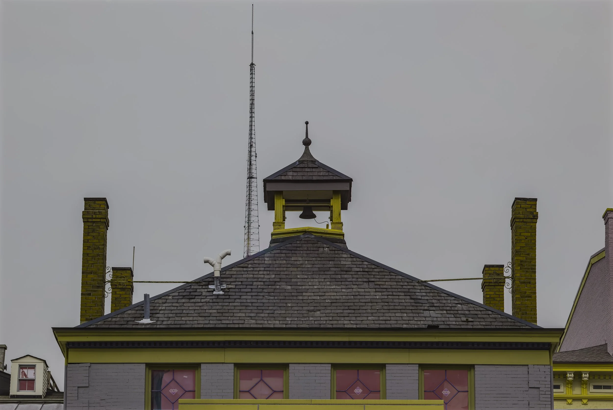 View of the top of a building with a small cupola, two yellow brick chimneys, and an antenna against a gray, overcast sky.