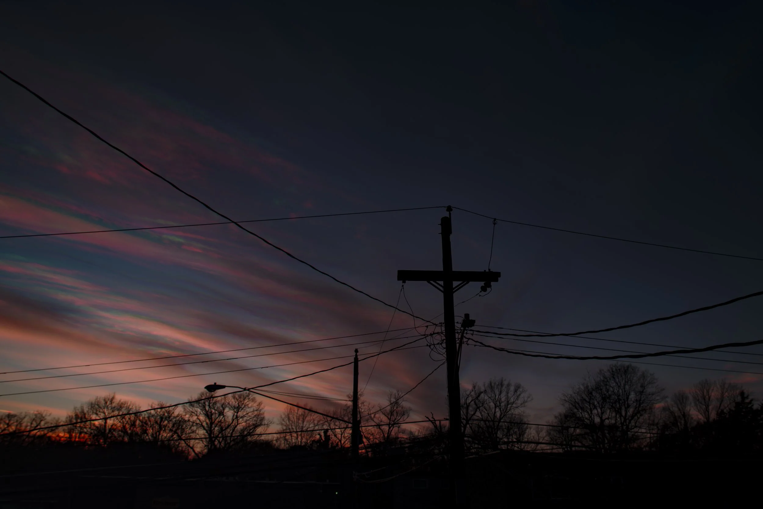 Silhouette of utility poles and wires against a colorful sunset or dawn sky with trees in the foreground.