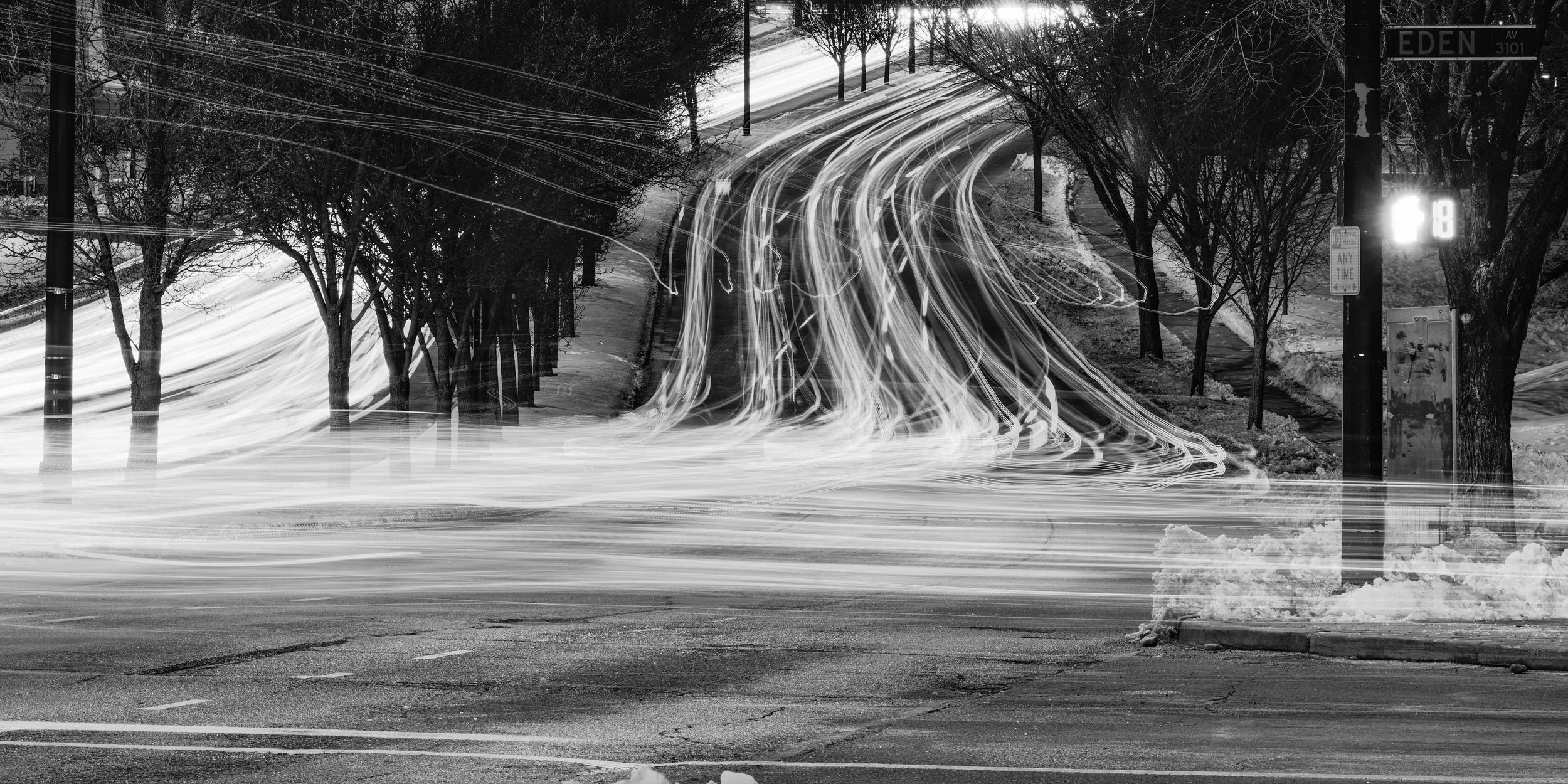 Nighttime black and white photo of a street with streaks of car lights indicating long exposure. Trees line the street, and there's a street sign reading 'Eden Ave' and a digital sign showing the number 8.