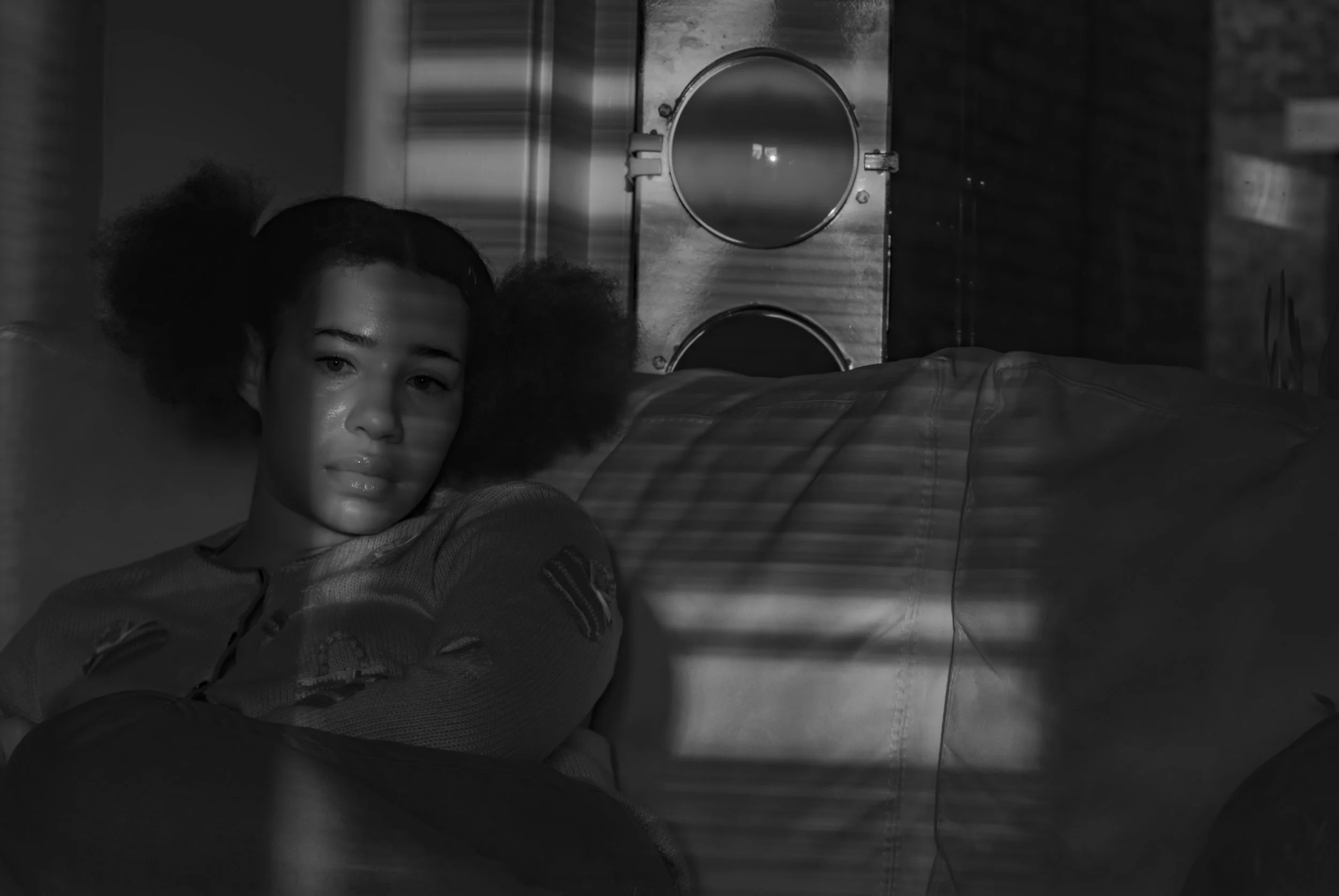 A young woman with dark hair in puffs sitting on a couch in a dimly lit room, with shadows from blinds cast across her face and the background, which includes a speaker and brick wall.