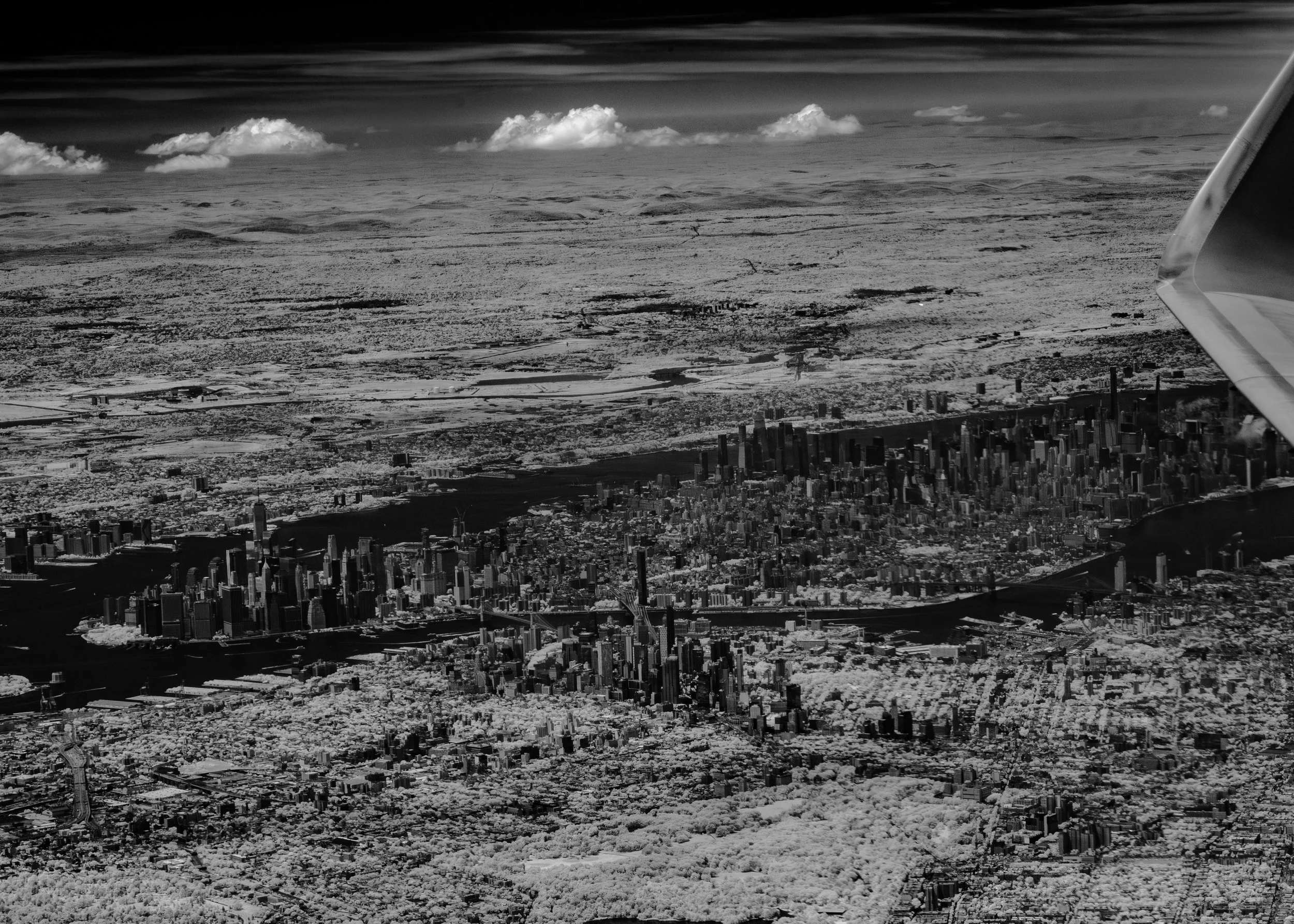 Aerial black-and-white view of New York City with skyscrapers, rivers, and surrounding landscape from an airplane window.