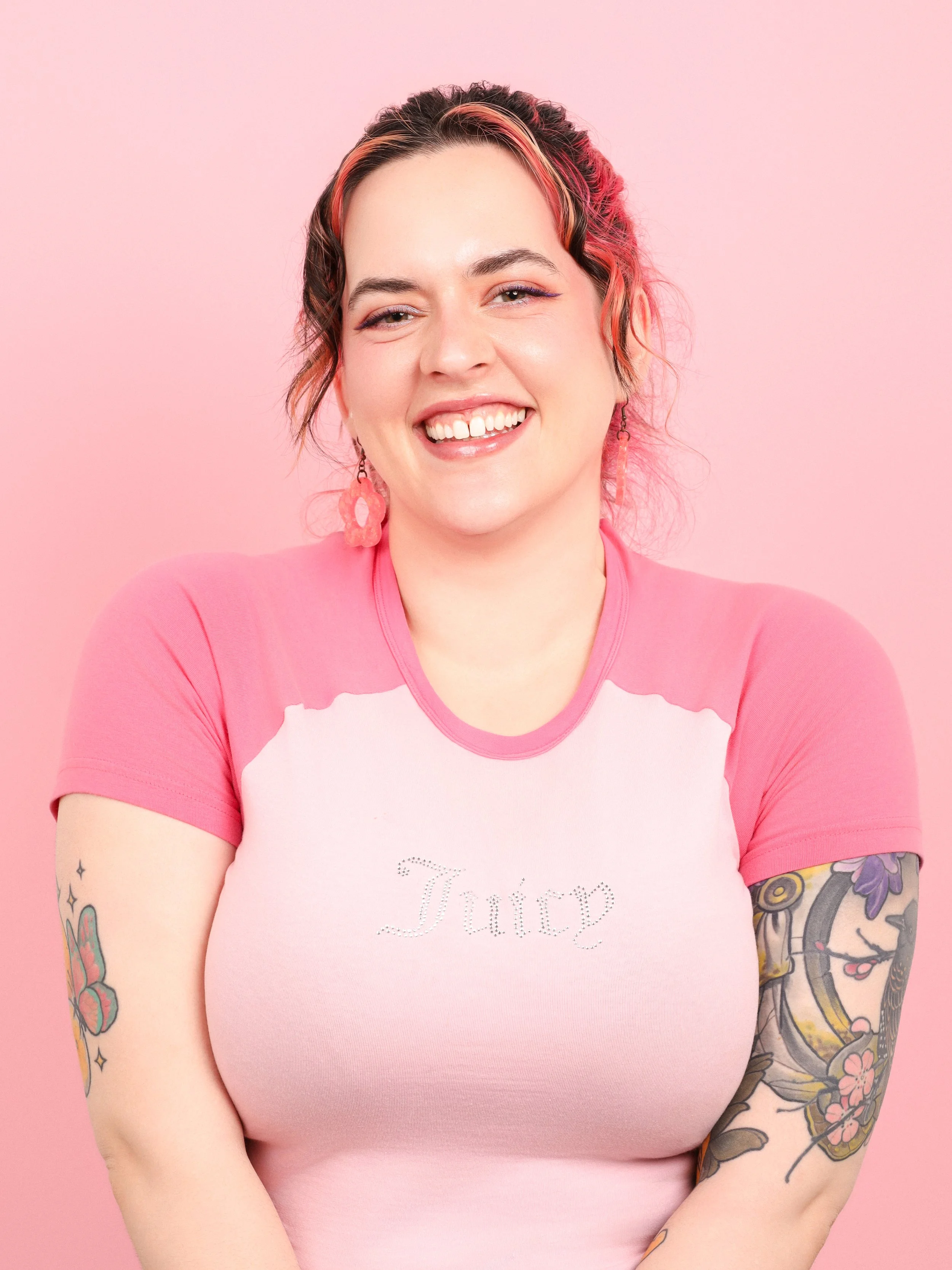 A while woman with pink and dark brown hair and tattoos on her arms looks at the camera smiling. she is wearing a pink T shirt.
