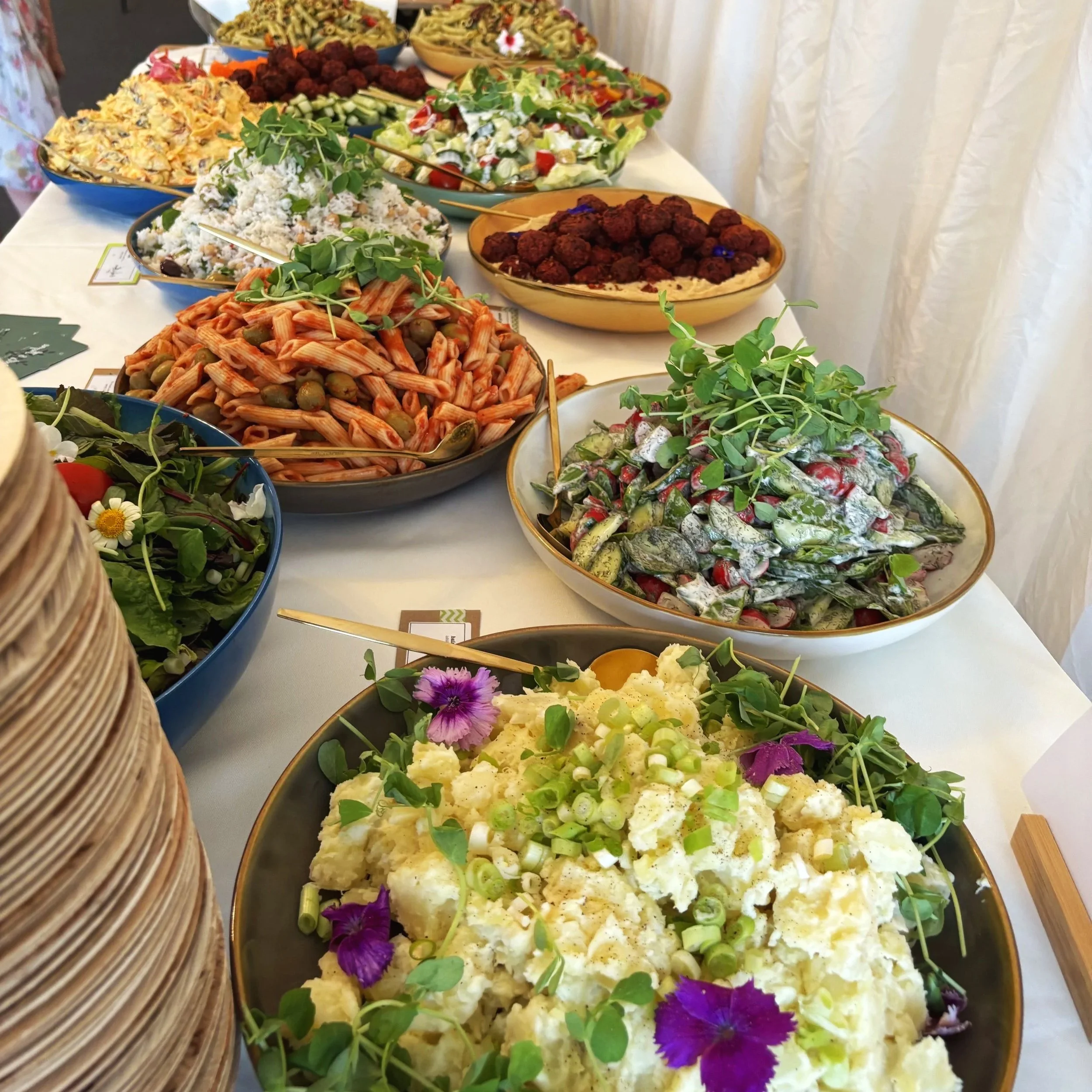 A variety of colorful salads and pasta dishes displayed on a long buffet table, decorated with fresh edible flowers and greens.