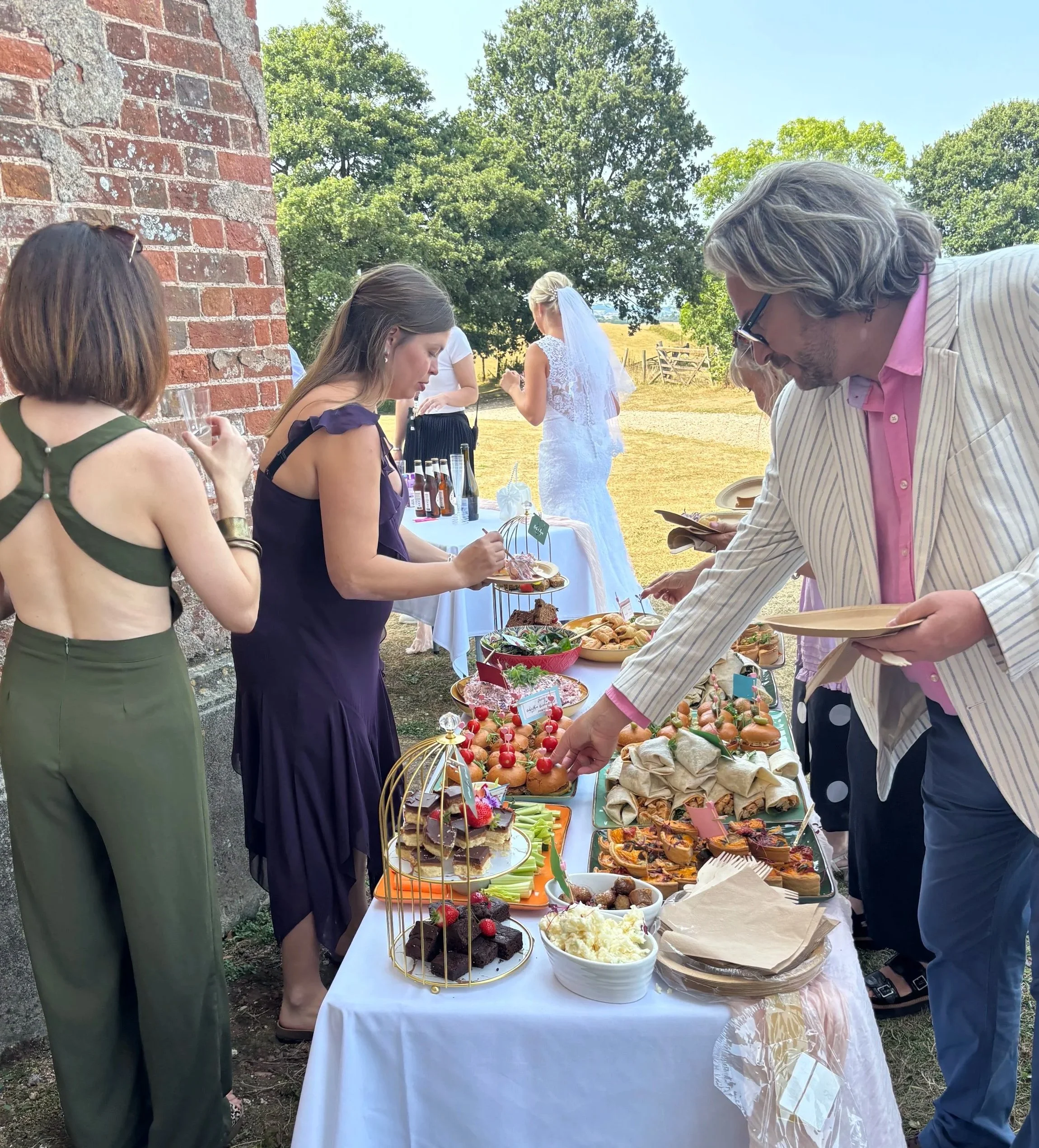 People attending a wedding reception outdoors, selecting from a table of desserts including mini cakes, fruit skewers, and pastries, in front of a brick building with green trees and a grassy field in the background.