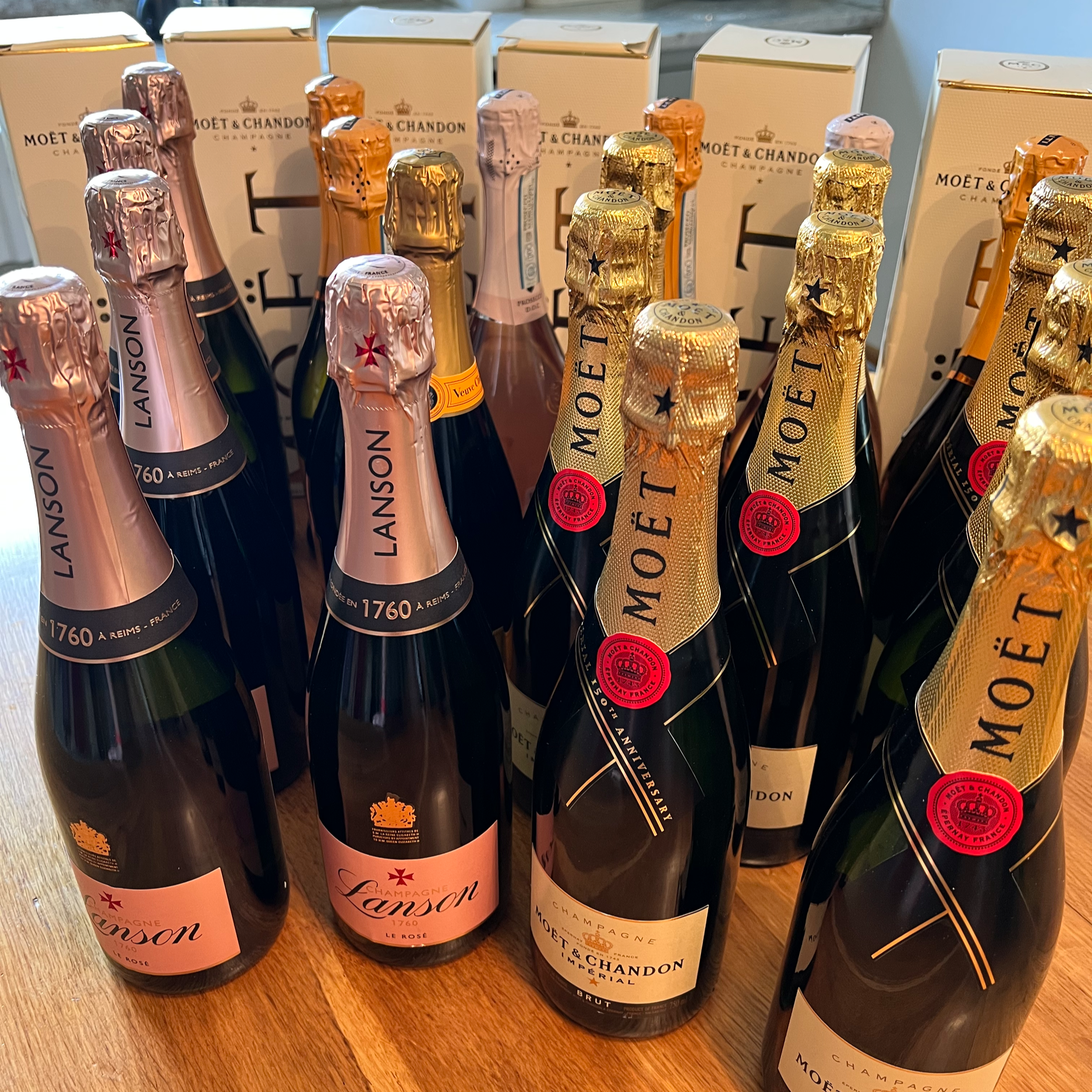 Multiple bottles of Moët & Chandon champagne with pink and gold foil caps, arranged on a wooden surface with boxes in the background.