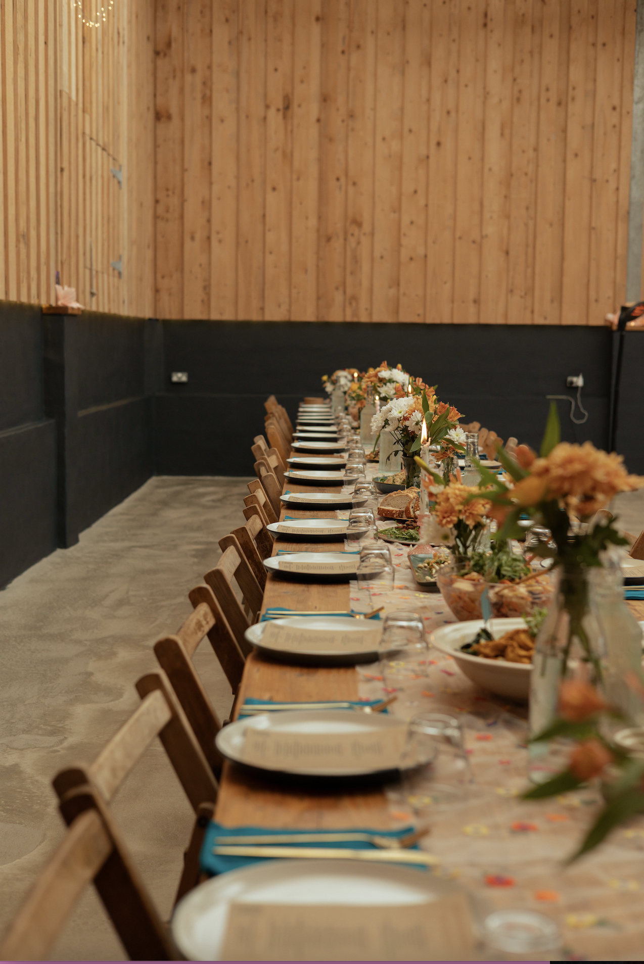 Long dining table set for a meal with floral centerpieces, dinner plates, glasses, napkins, and utensils in a rustic wooden room.